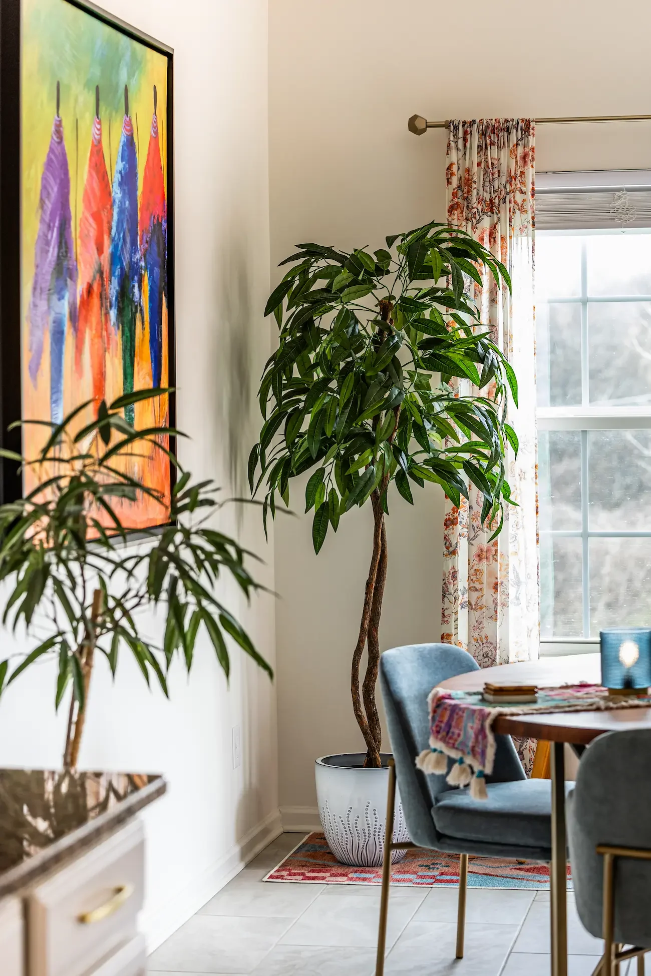 A cozy dining area with a large potted plant near a window with floral curtains, colorful tablecloth, and a partial view of a round table and chairs.