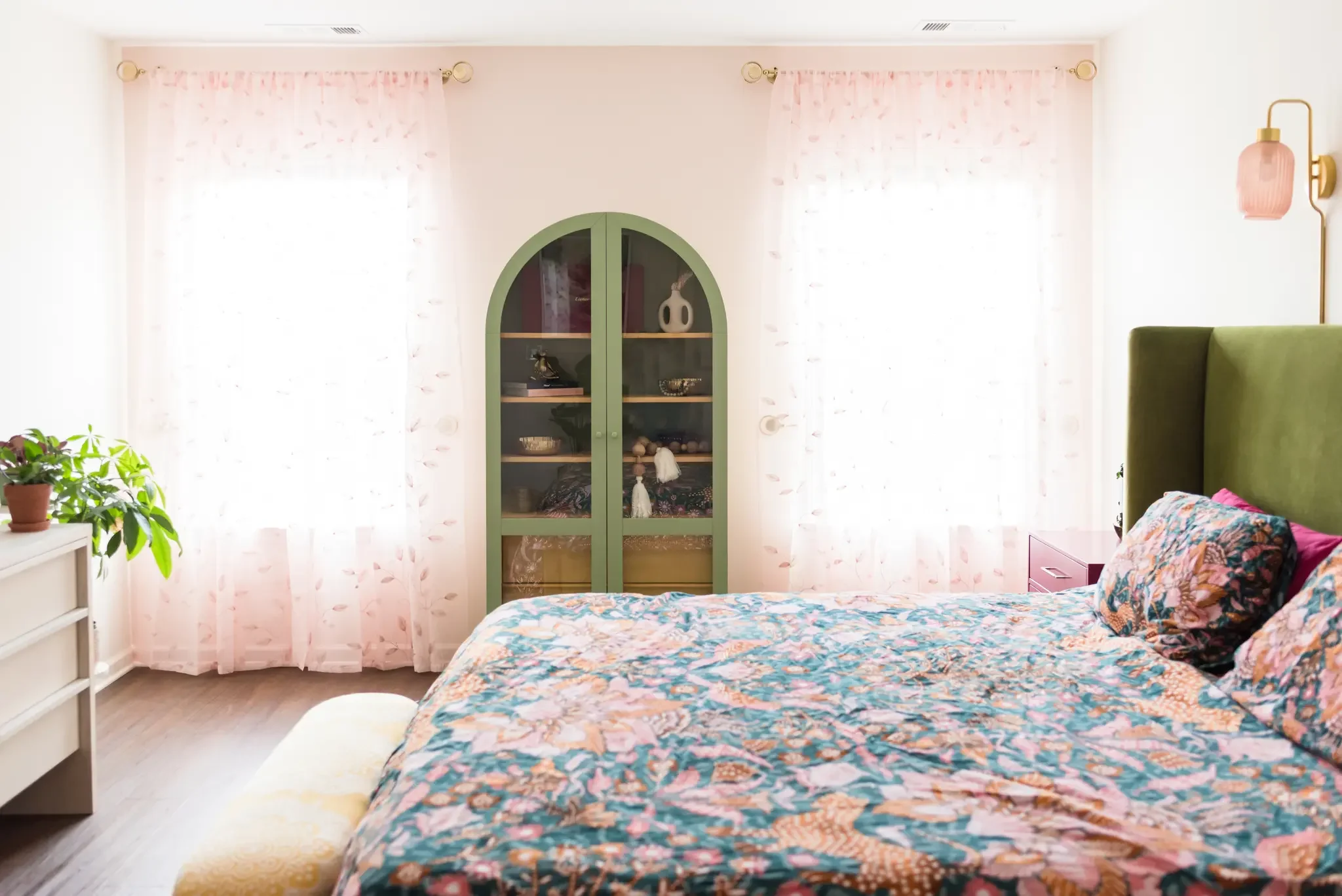 Bright bedroom with a large bed covered in a colorful floral quilt, green headboard, pink nightstand, pink wall-mounted lamp, white curtain with pink leaf pattern, arched green display cabinet, and a potted plant on a white dresser.