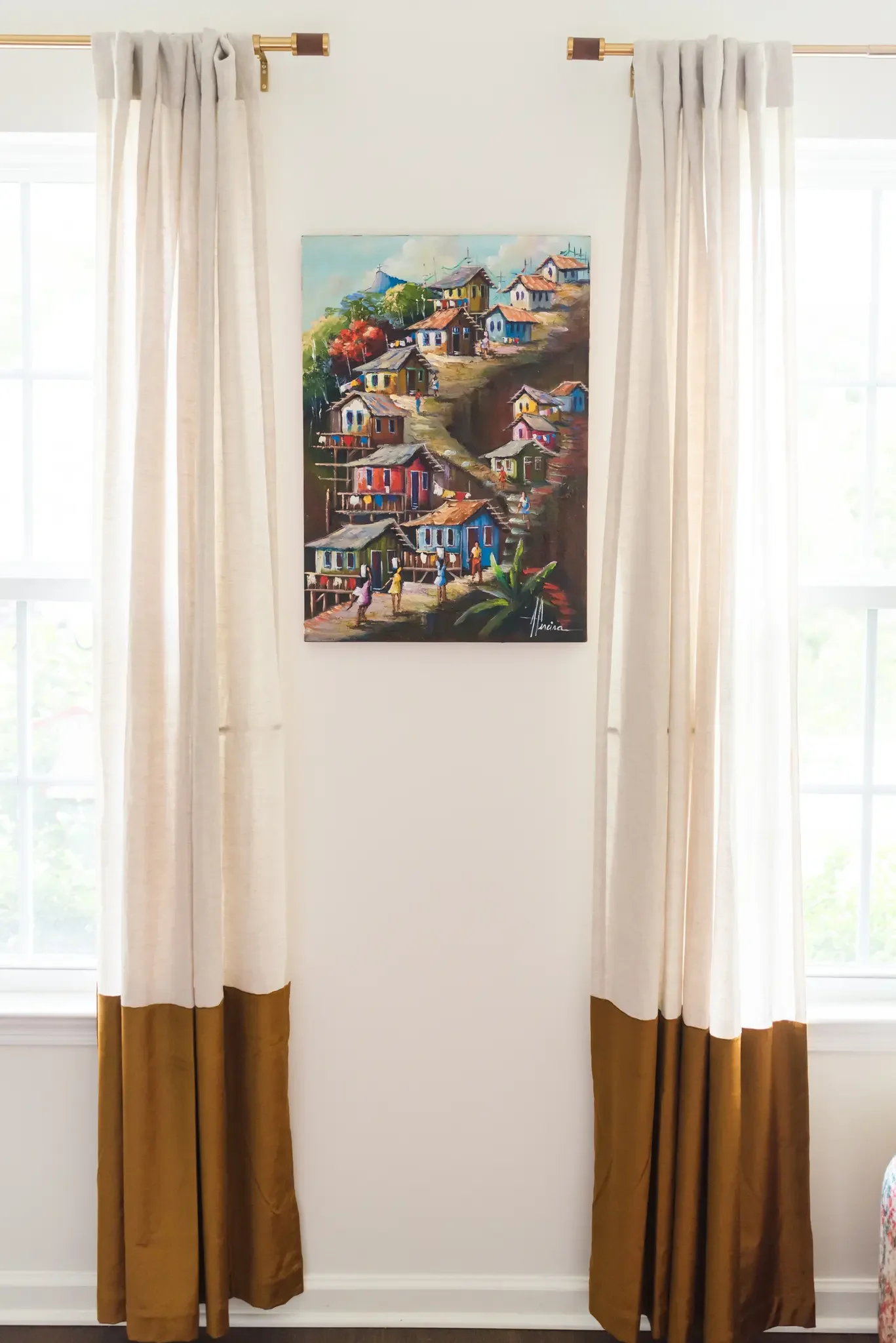 Painting of colorful houses on a hillside between two windows with white curtains.