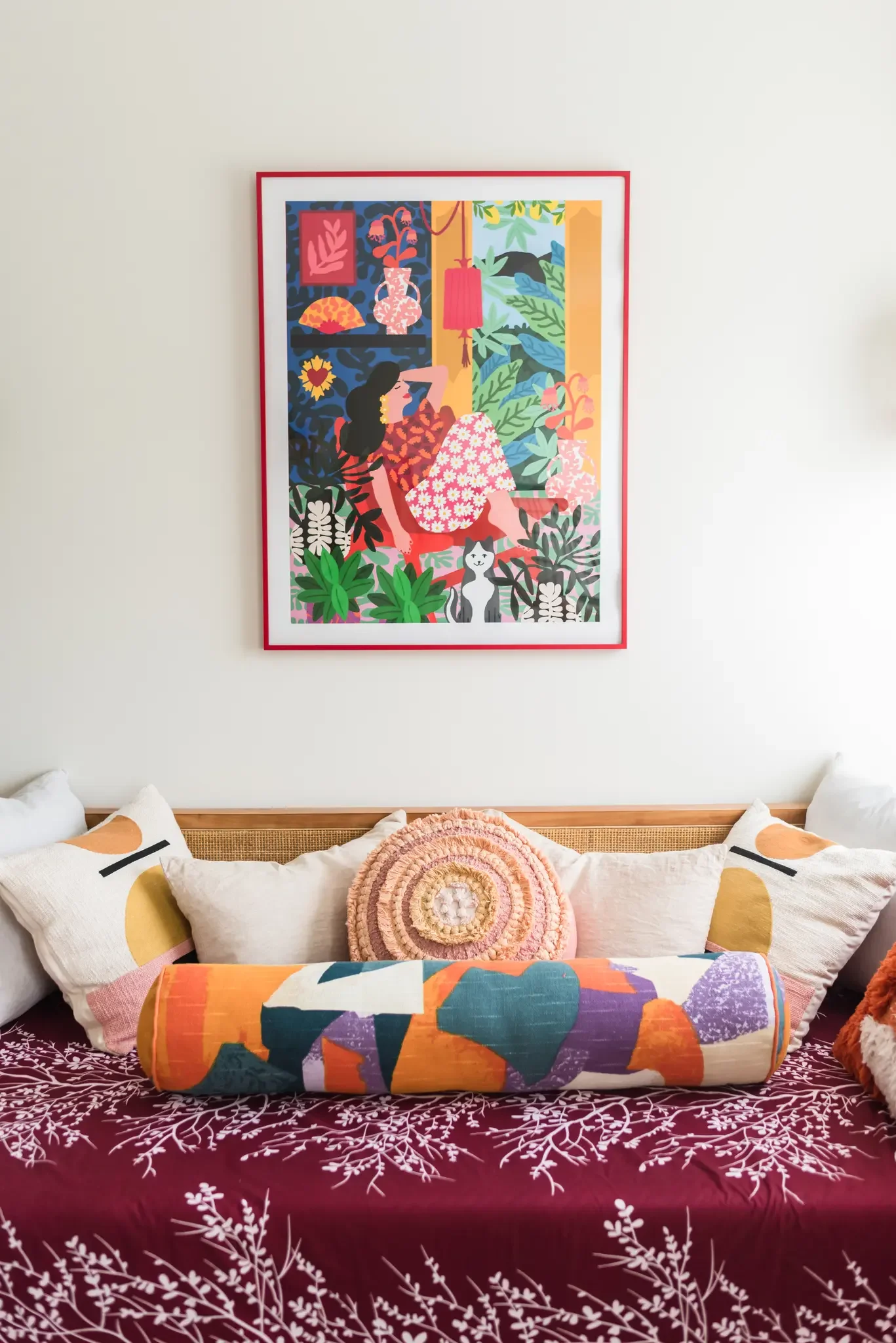 Living room with a bed, multiple decorative pillows, vibrant purple bedspread with white leaf pattern, colorful art print on wall, and a framed illustration on top of a shelf, surrounded by lush plants.