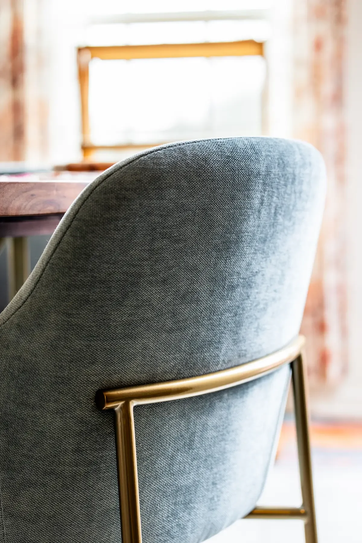 Close-up of a grey upholstered chair with gold metal armrest, in front of a window with curtains and a wooden table, bright natural light.