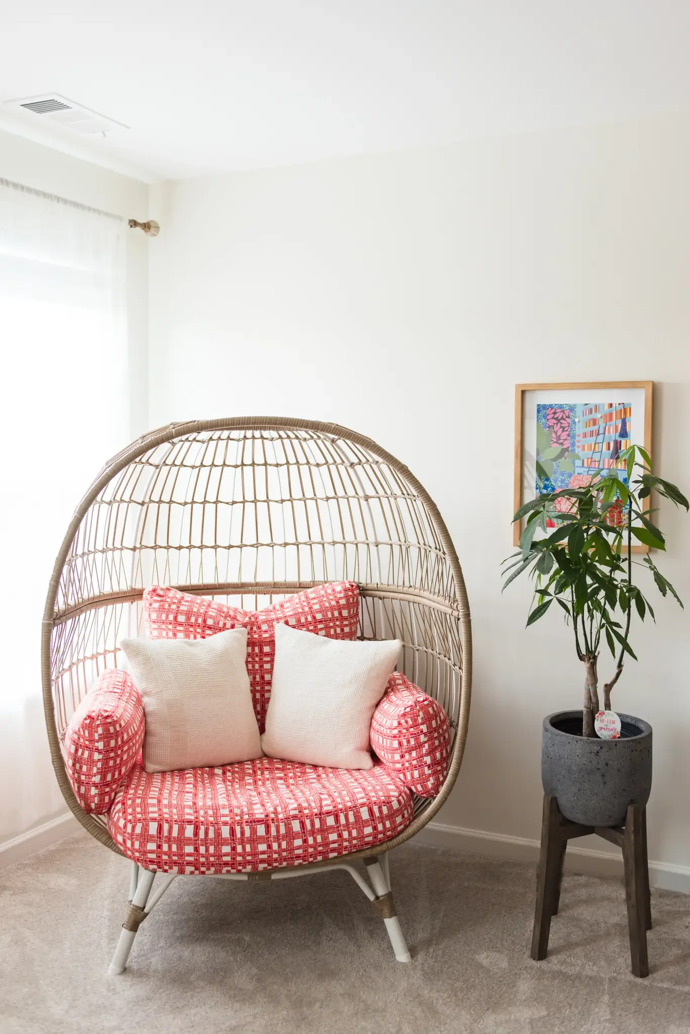 A cozy indoor space featuring a round wicker chair with red and white checkered cushions and white pillows, a wooden side table with a potted plant, a framed colorful artwork on the wall, white curtains, and a light-colored carpeted floor.