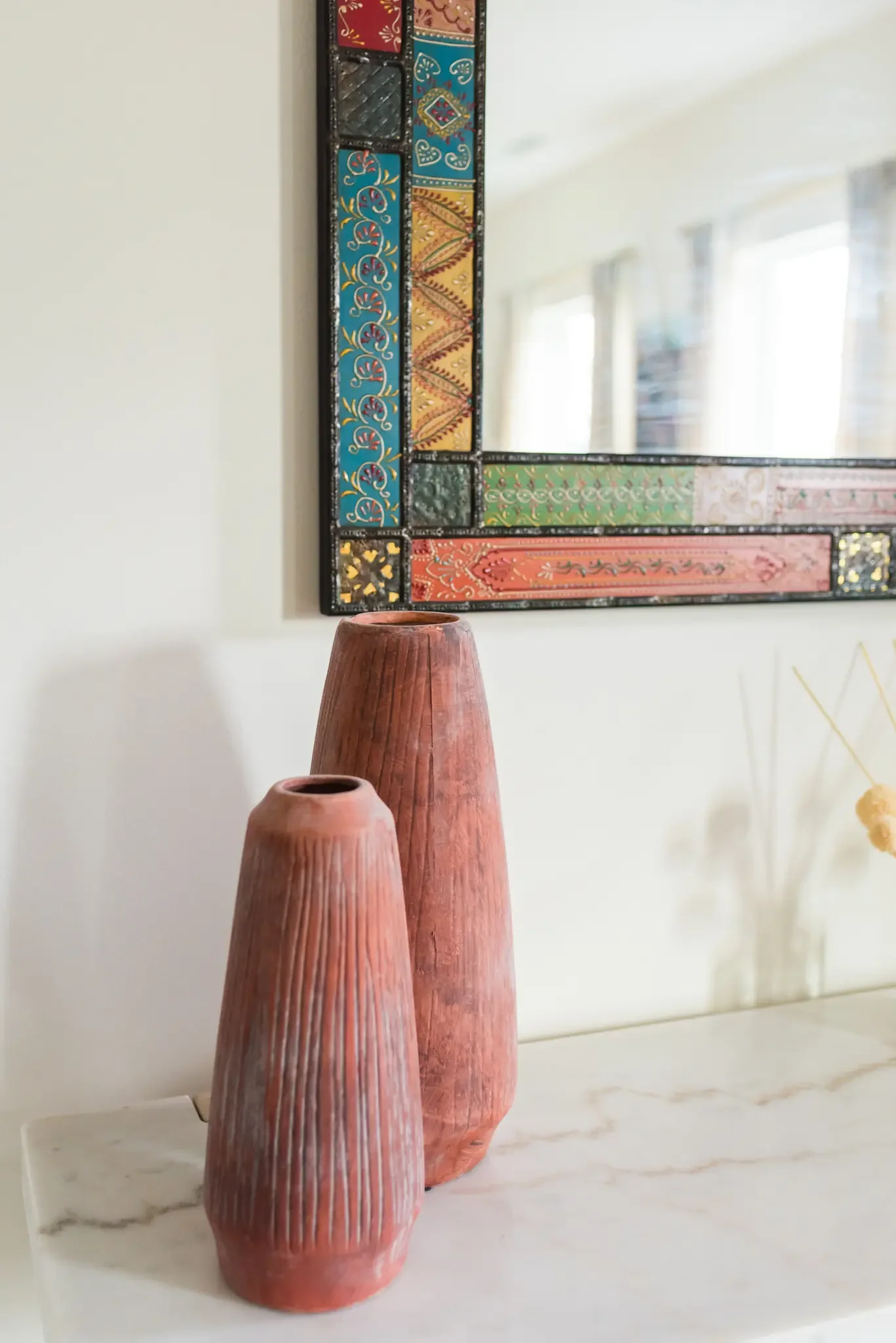 Three pinkish-orange ceramic vases of varying heights on a white marble surface, with a colorful decorative mirror hanging on a white wall in the background.