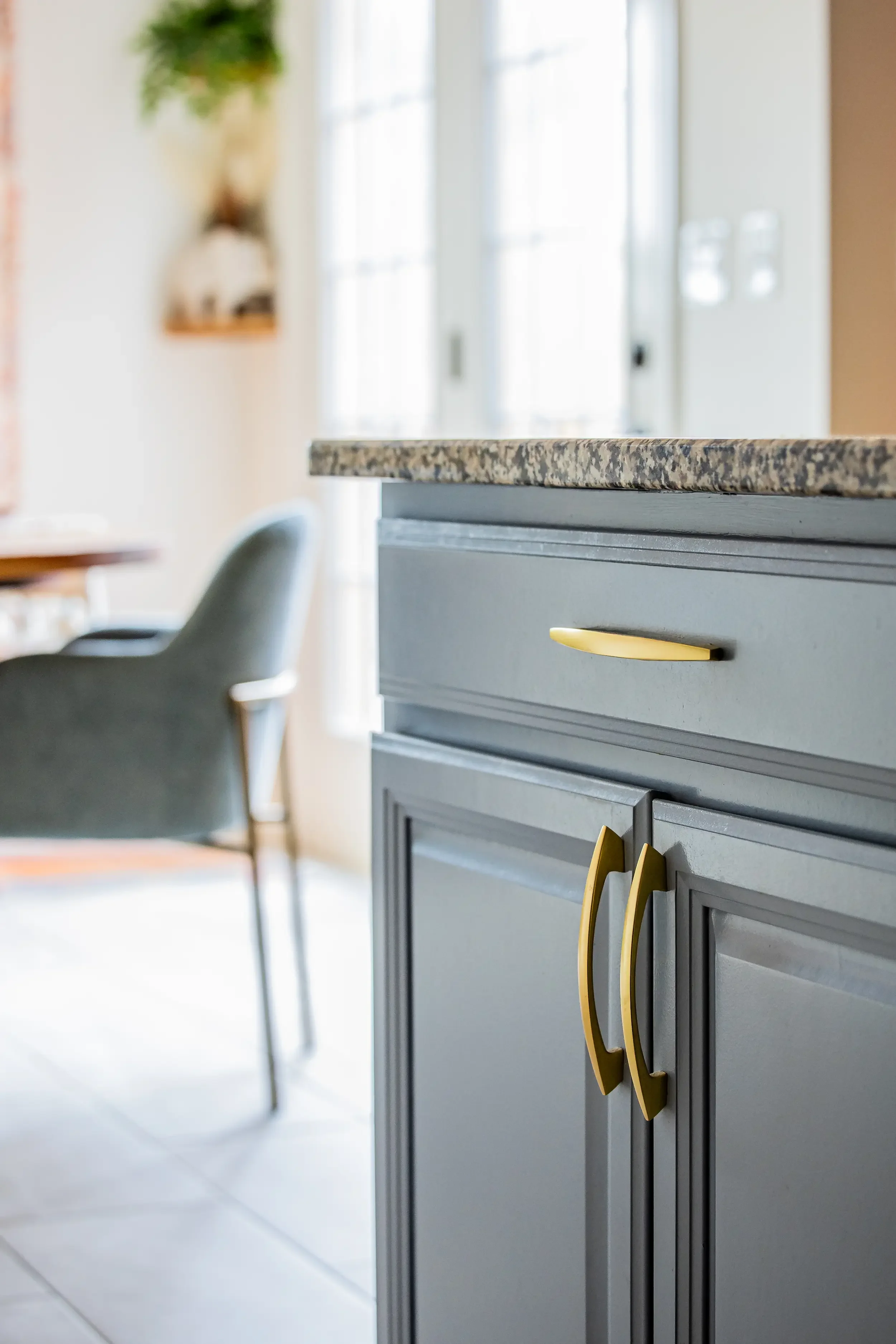 Close-up of a gray cabinet with yellow handles in a bright, modern kitchen, with a granite countertop and a blurred dining area with a chair in the background.
