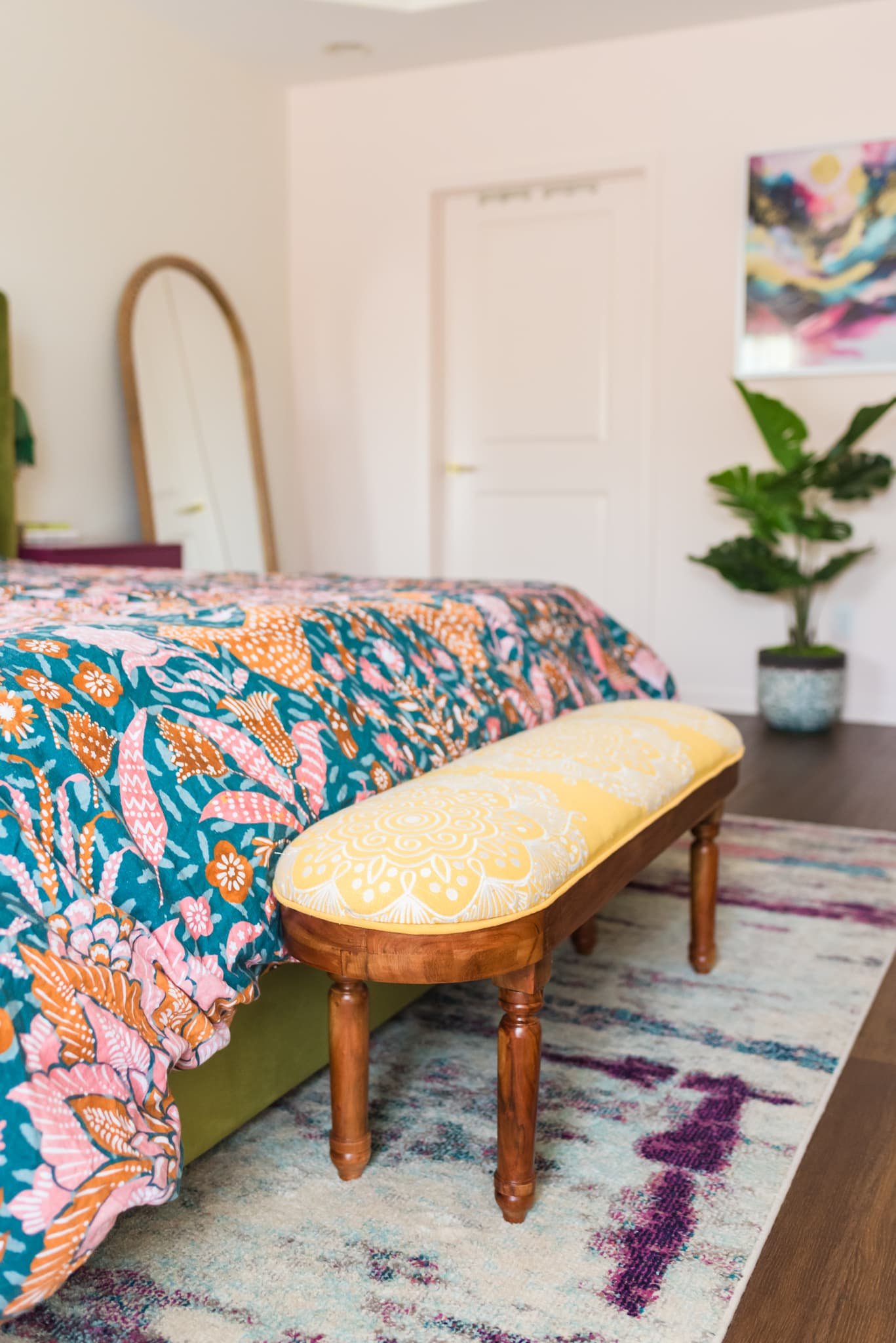 A bedroom with a colorful floral bedspread, a wooden bench with a yellow cushion, a large green plant in a pot, a wall mirror, a painting, and a closed white door.