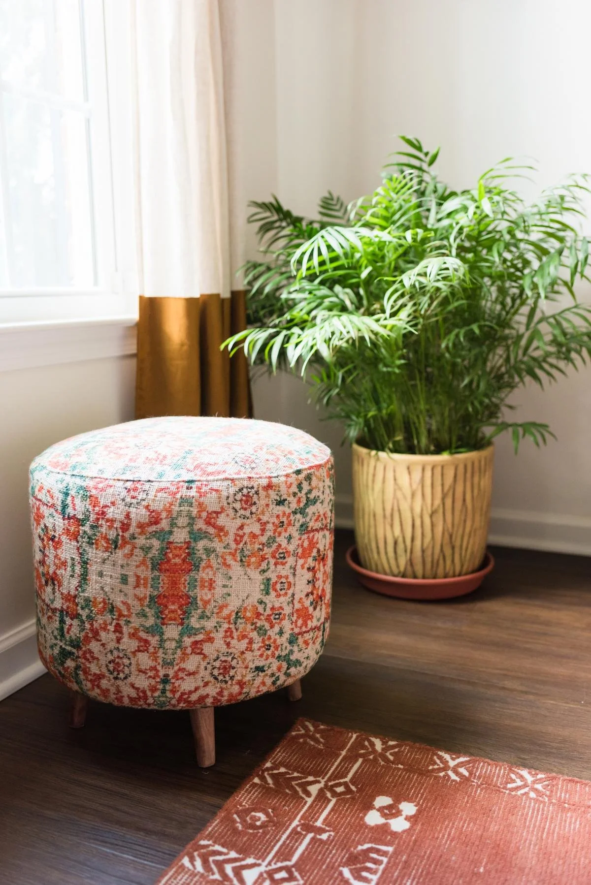 A colorful upholstered stool with four wooden legs, placed on a dark hardwood floor next to an orange patterned area rug. Behind the stool is a large green houseplant in a decorative yellow pot with a pink saucer, near a white wall with a window that has white curtains with a brown border.