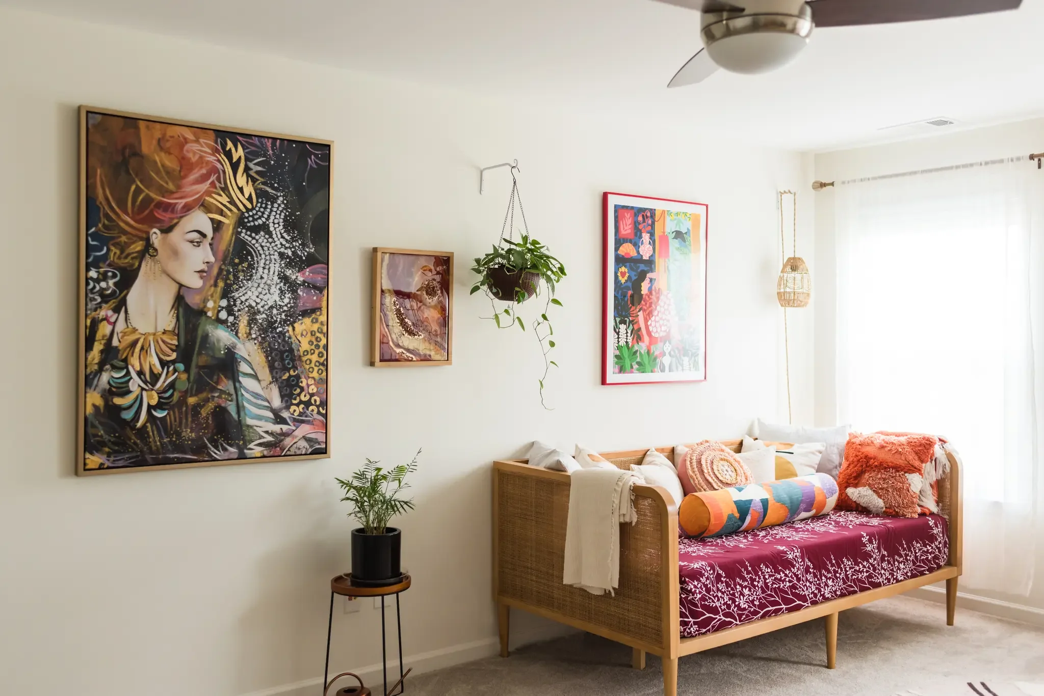 Living room with colorful artwork on the walls, a hanging plant, a wooden sofa with patterned and pink throw pillows, and a wicker pendant light. A small side table with a potted plant is nearby.
