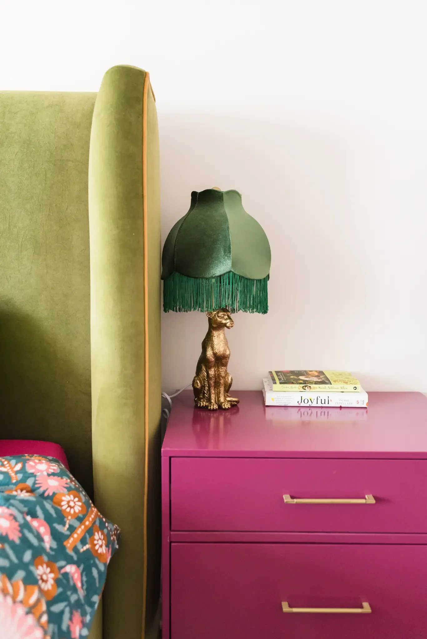 A green headboard bed with floral-patterned bedding, a purple dresser with two pulls, a green lamp with fringes and a dog figurine on top, and a stack of three books including 'Joyful'.