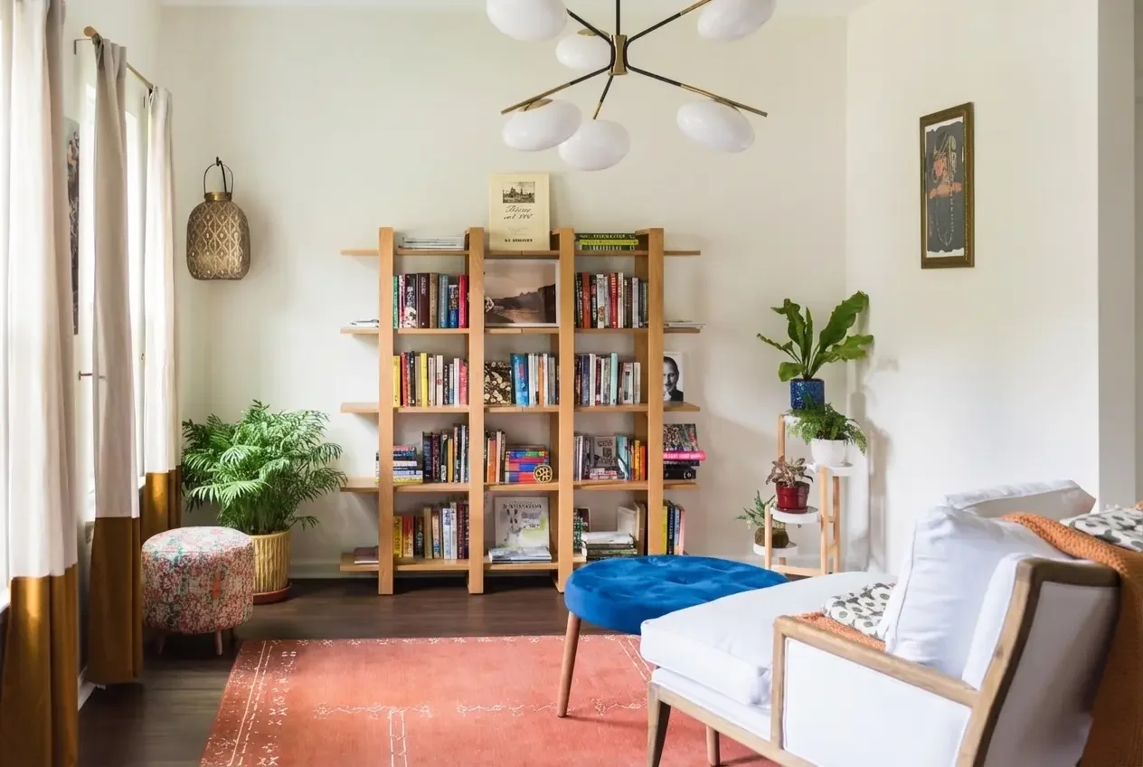 Living room with a wooden bookshelf filled with books, a blue round ottoman, a white couch with patterned cushions to the right, potted plants, hanging light fixture with multiple bulbs, curtains on the window, and a framed picture on the wall.
