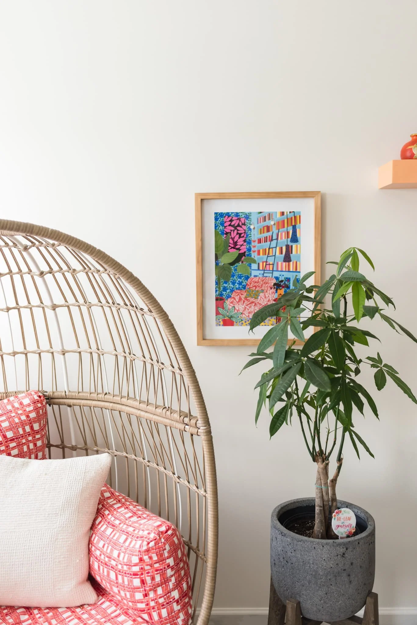 A cozy corner of a room featuring a wooden rattan-style chair with red patterned cushions and pillows. Beside the chair is a tall potted plant with broad green leaves in a gray concrete pot, standing on wooden legs. On the wall behind, there is a colorful framed artwork and a small orange shelf with a decorative red vase.