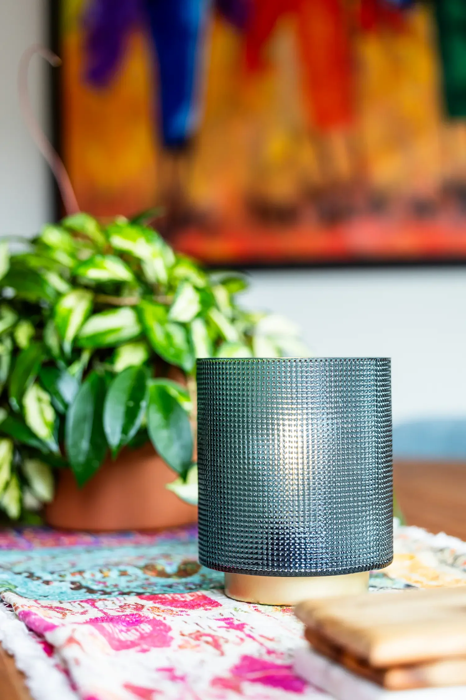 A black textured candle holder on a table with colorful textiles, a potted leafy plant, and blurred wall art in the background.