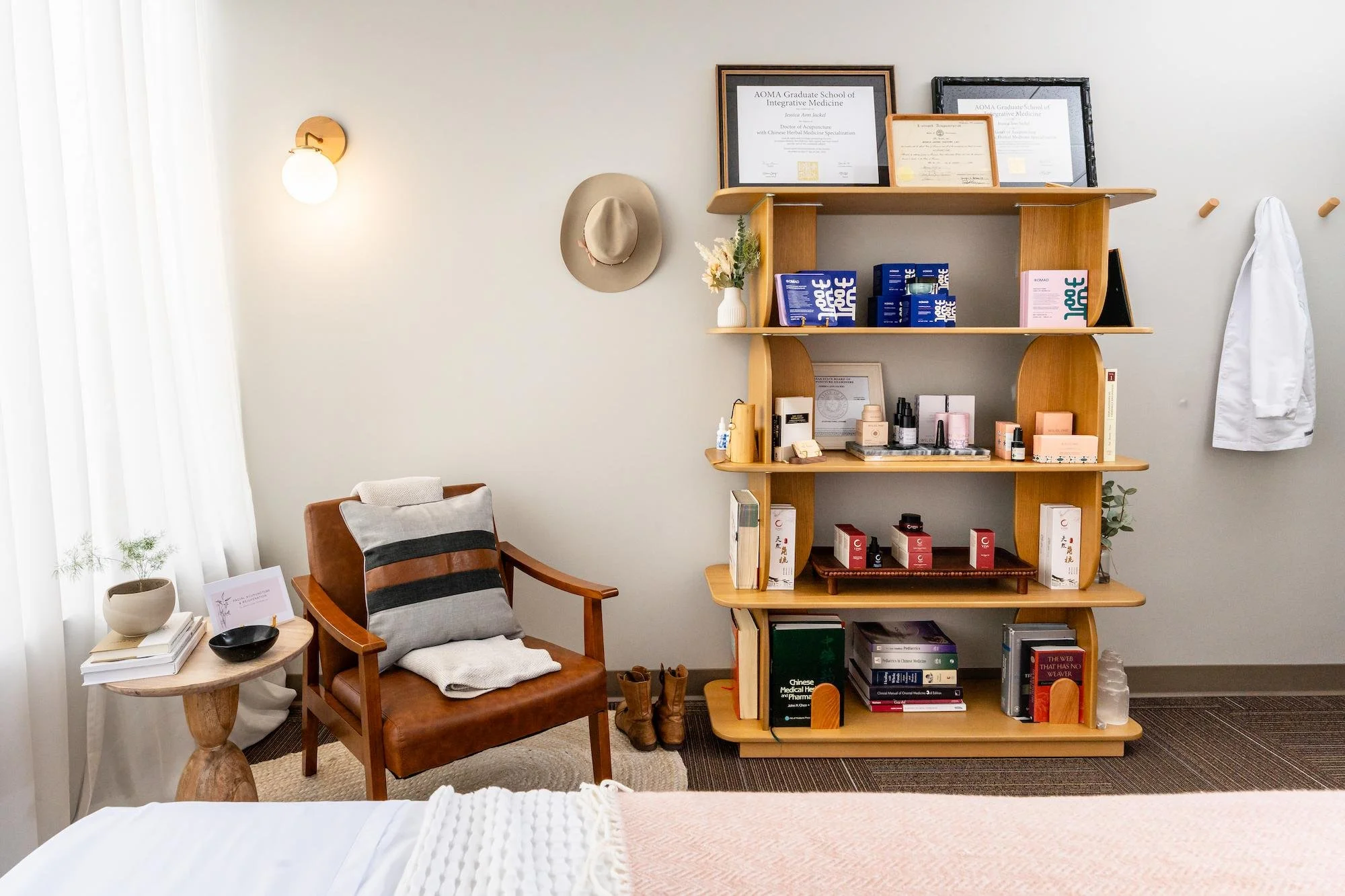 A cozy room corner with a wooden armchair, a round side table with stacked books and a plant, a tall wooden bookshelf filled with books and framed certificates, and a wall-mounted light fixture, creating a warm and inviting atmosphere.
