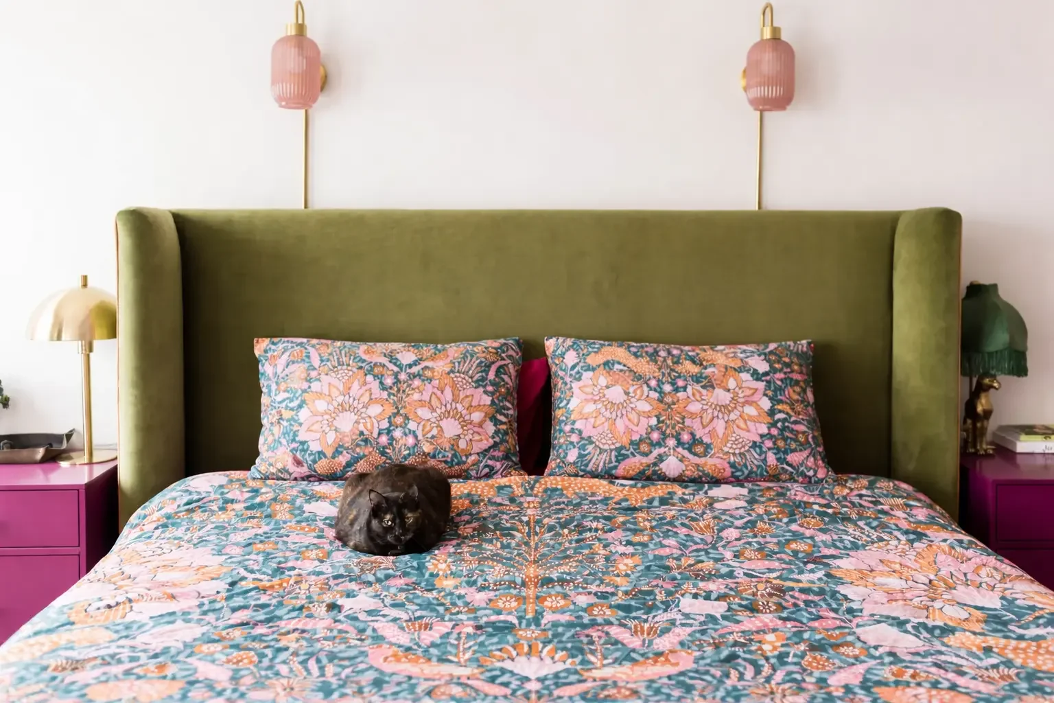 A bedroom with a large green headboard, floral bedding, and two matching floral pillows. There are pink and green lamps on either side of the bed, and a black cat lying on the bed.