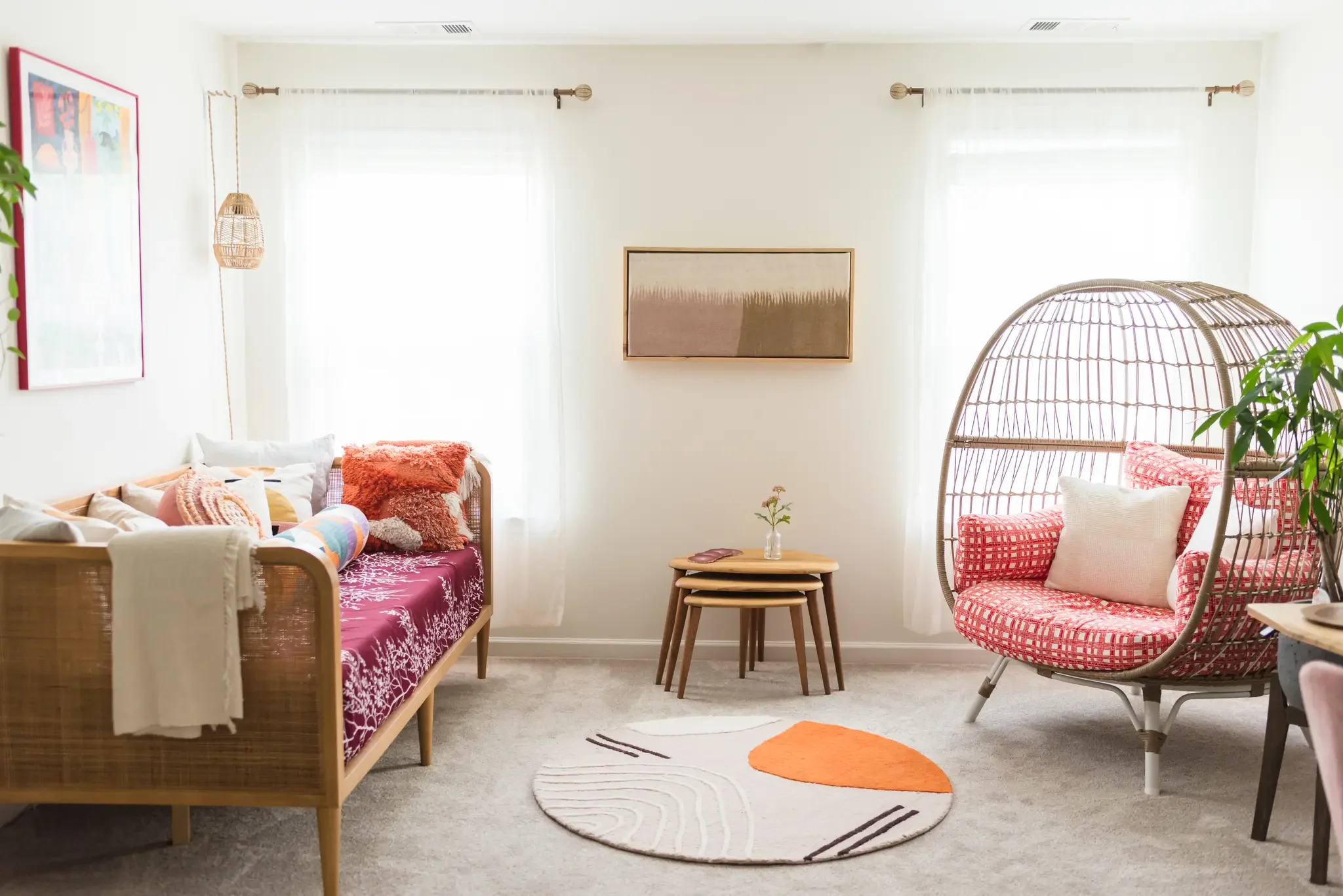 A cozy living room with a wooden daybed on the left decorated with colorful pillows and a pink blanket, a small wooden side table in the center with a small plant, and a rattan hanging chair on the right with cushions and throw pillows. The room has large windows with white curtains, candle-like curtain rods, a wall art piece, and a modern patterned rug on the carpeted floor.