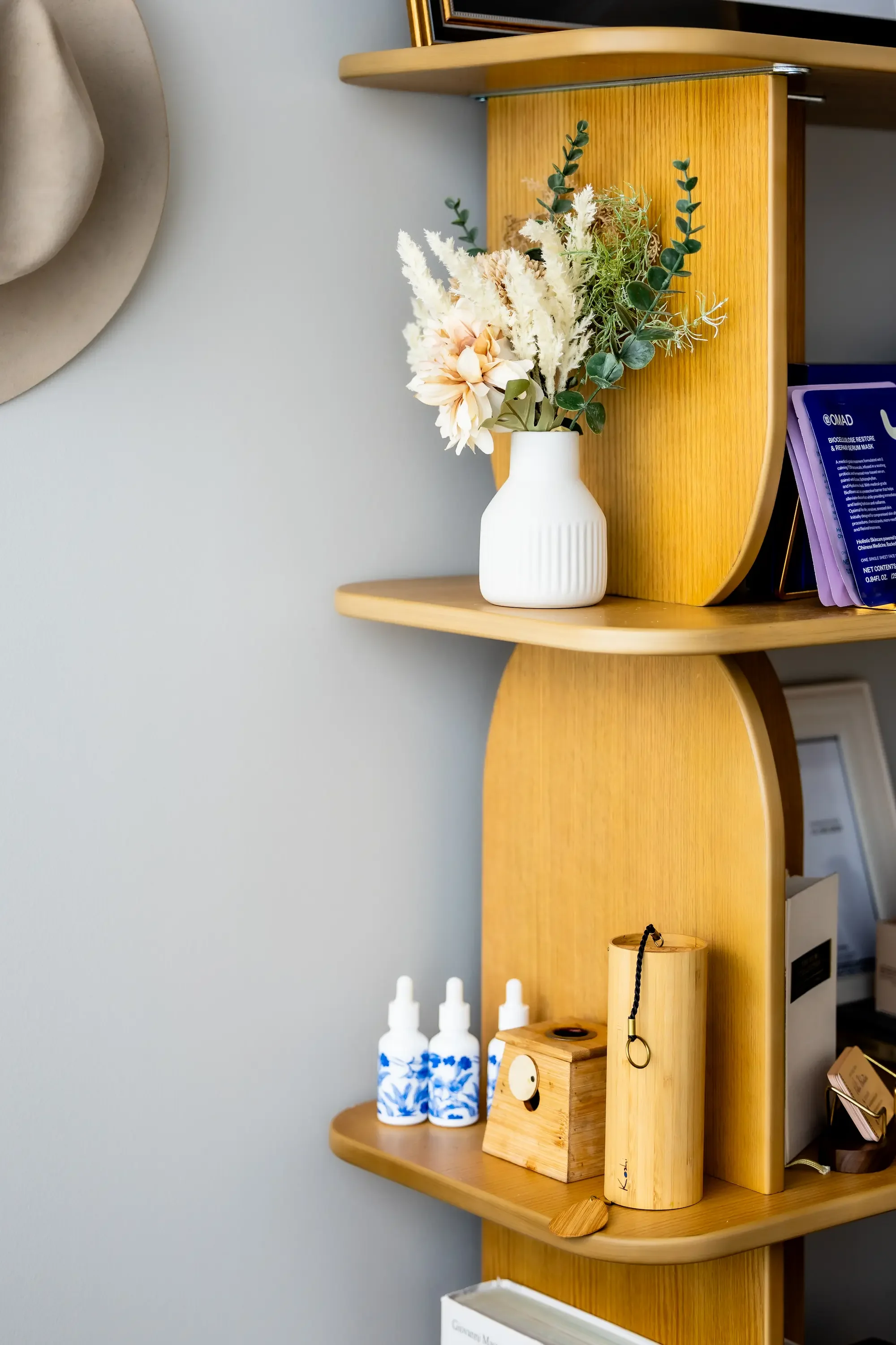 A wooden shelf with a white vase holding dried flowers and green foliage. On the shelf below, there are three white bottles with blue designs, and a wooden box with a round, dark element on top. Part of another wooden object and some bottles are visible at the bottom.