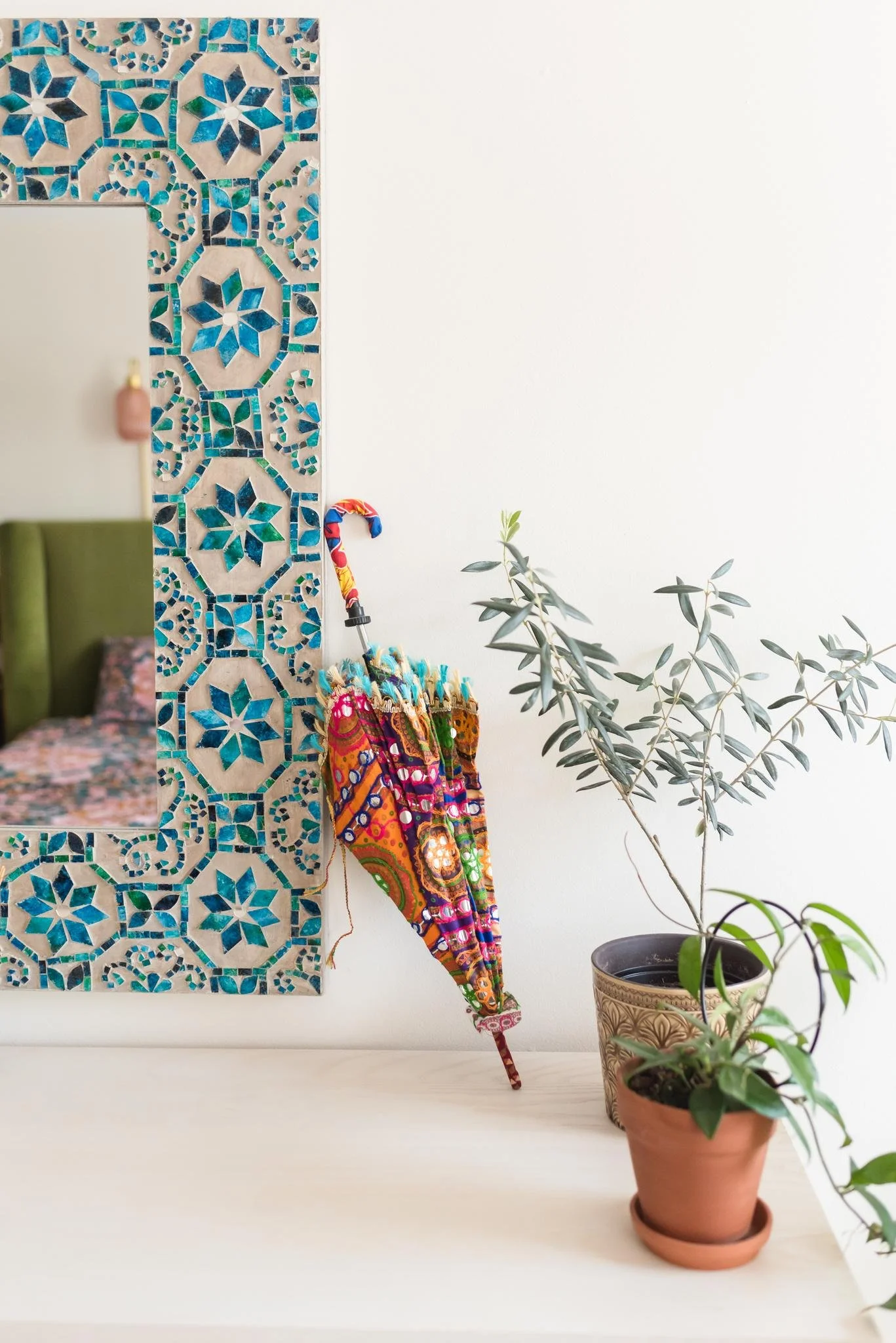 Interior decor featuring a mosaic tile mirror with blue and green patterns, colorful fabric umbrellas, and potted green plants on a white surface.