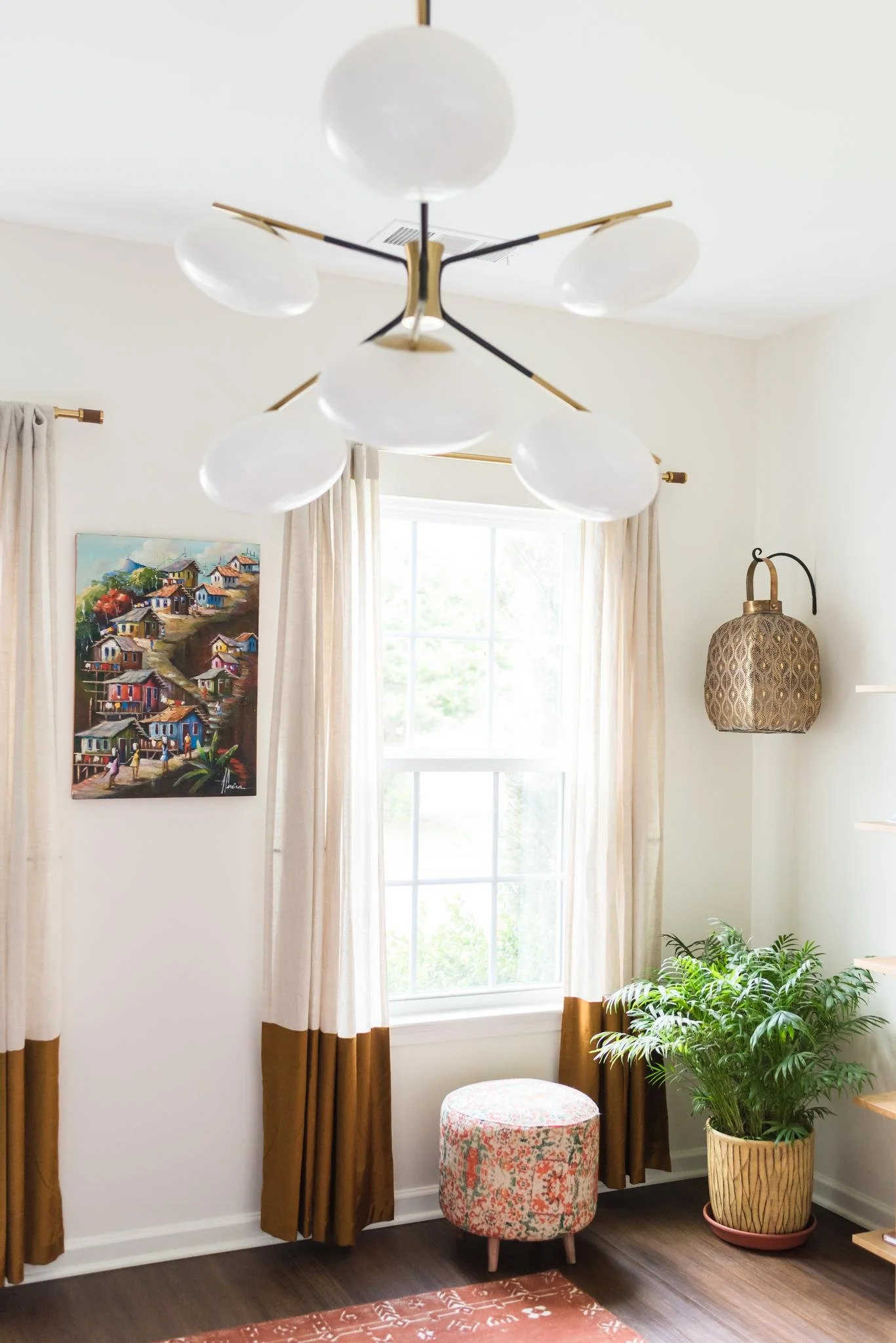A cozy interior corner with a large window with white trim, beige curtains with brown bottom borders, a colorful floral ottoman, a lush green potted plant, and a decorative wall lamp.