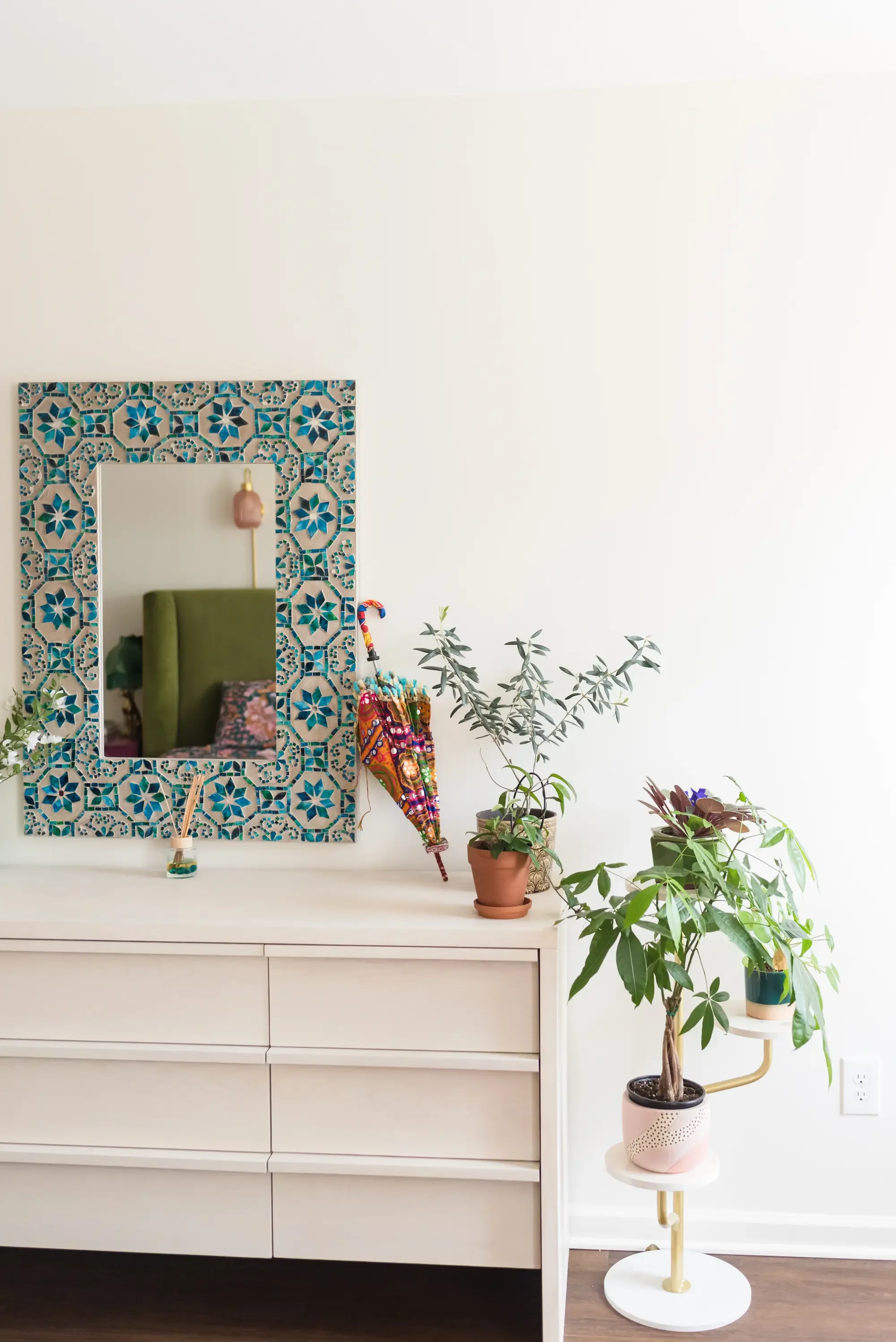 A modern bedroom dresser with plants on top and a decorative mirror with blue tile frame reflecting a green chair and bed with floral sheets.