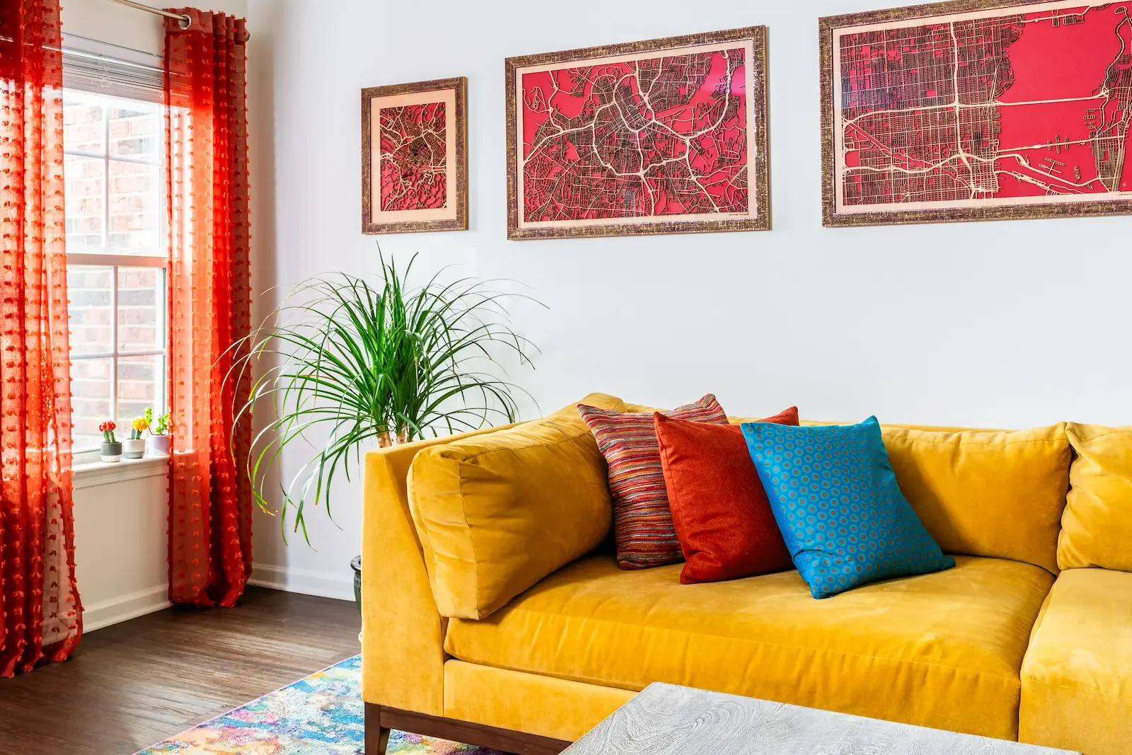 Living room with yellow sofa, colorful pillows, and framed city maps on white wall. Bright window with orange curtains, potted plants on windowsill, and hardwood floor.