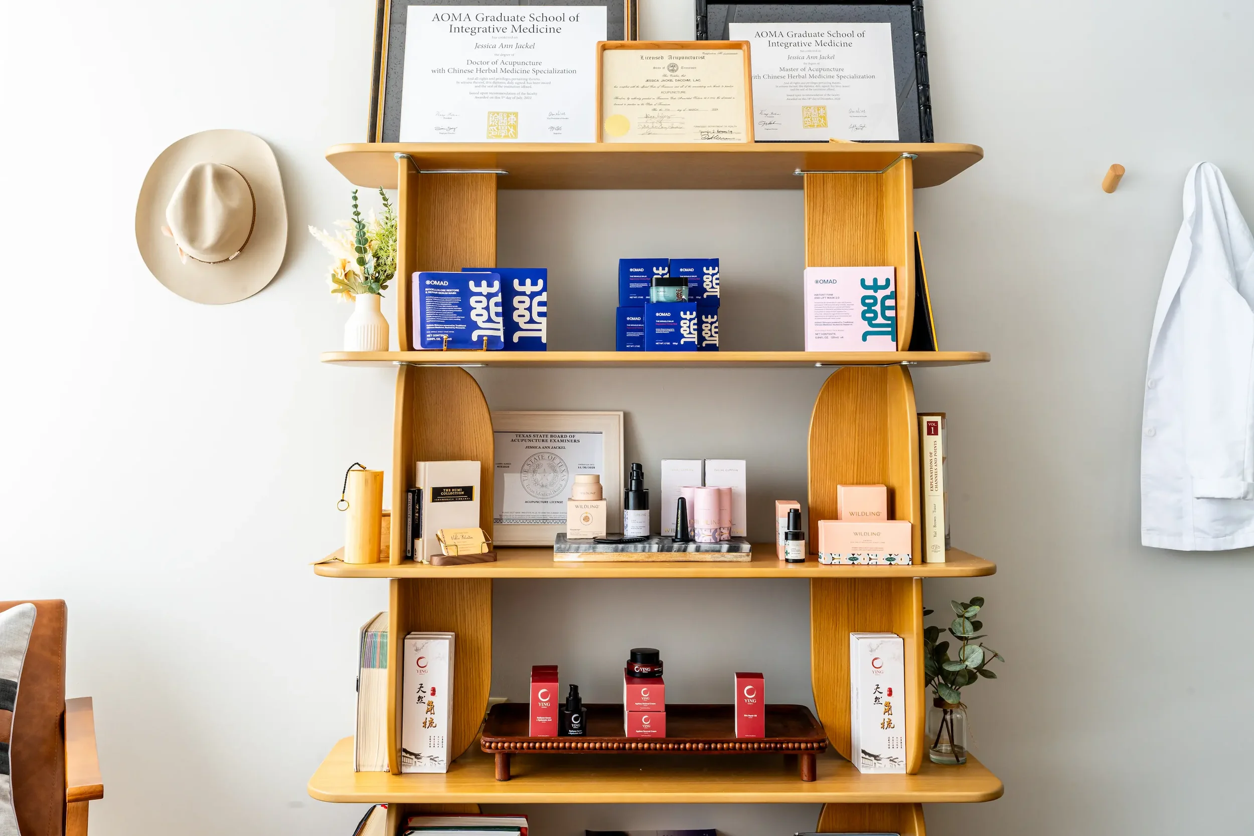 Wooden shelf with framed diplomas and awards at the top, a white vase with flowers on the second shelf, skincare and cosmetic products on the third shelf, and various boxes and bottles on the bottom shelf. A white coat hangs on a hook to the right, and a beige hat hangs on the wall to the left.