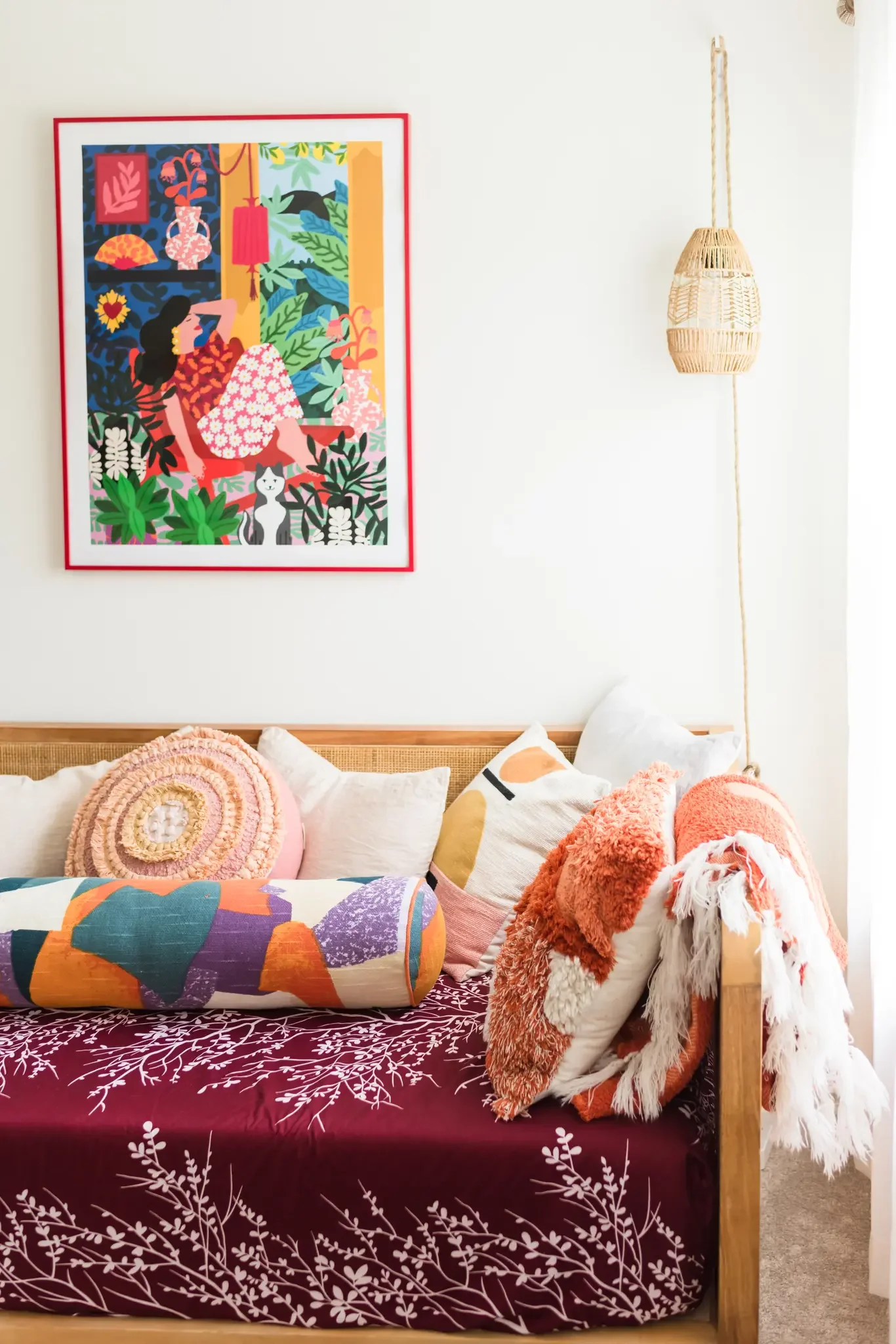 A bed with a maroon bedspread featuring white tree branches and leaves. There are multiple pillows including a round pink textured pillow, a white pillow with pink and orange design, and more. On the bed, there is a large, colorful bolster pillow with a geometric pattern. The wall behind the bed has a large colorful artwork featuring a woman sitting among lush green plants, a cat, and decorative objects. To the right of the artwork, a woven hanging lamp is suspended from the ceiling.