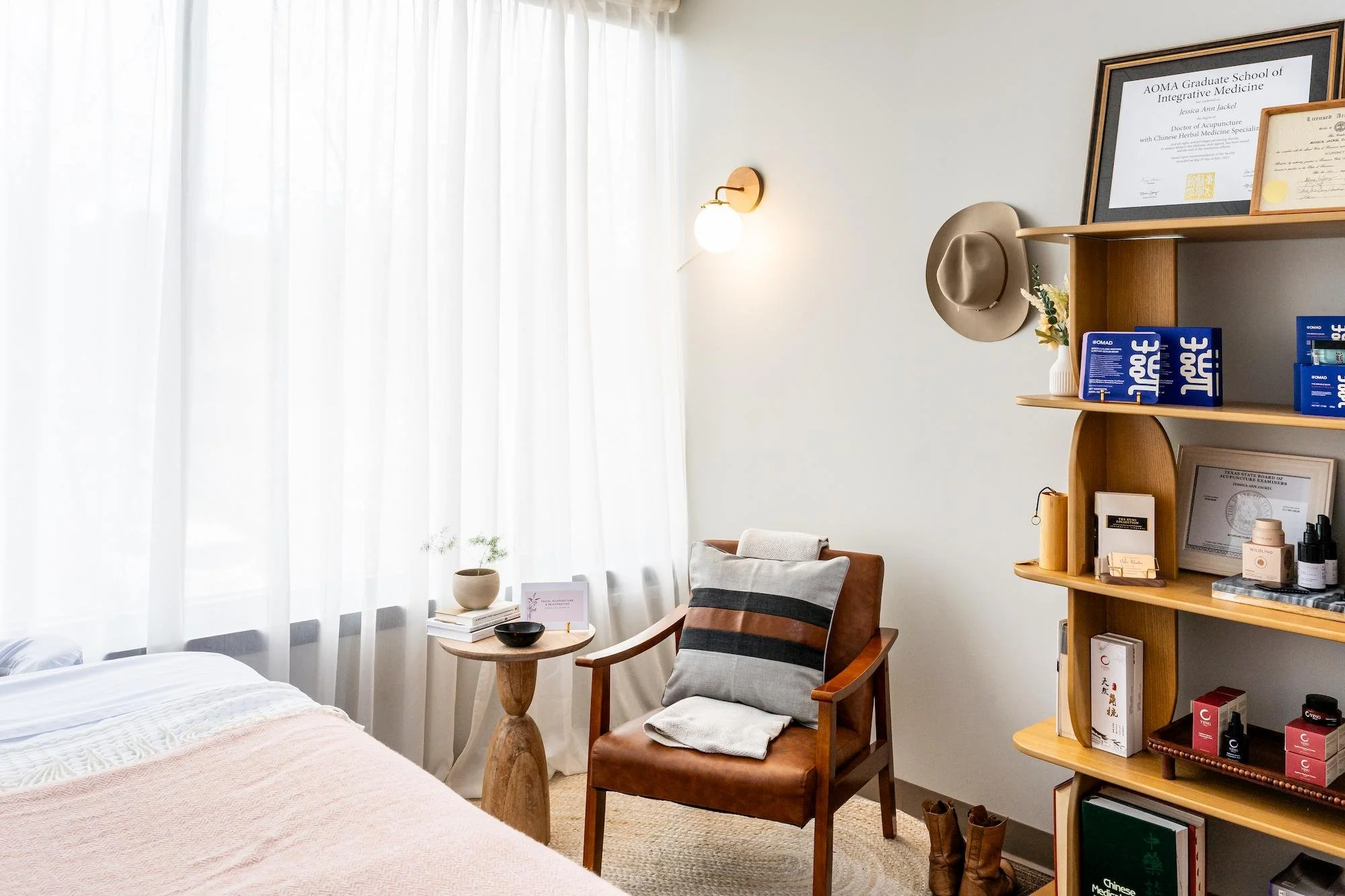 A cozy room corner with a bed, a wooden side table, a brown leather chair with a cushion, and a wooden bookshelf filled with books and small decorative items. The wall is decorated with framed certificates, a beige hat, and a wall-mounted light. The room has large windows with white sheer curtains allowing natural light to fill the space.