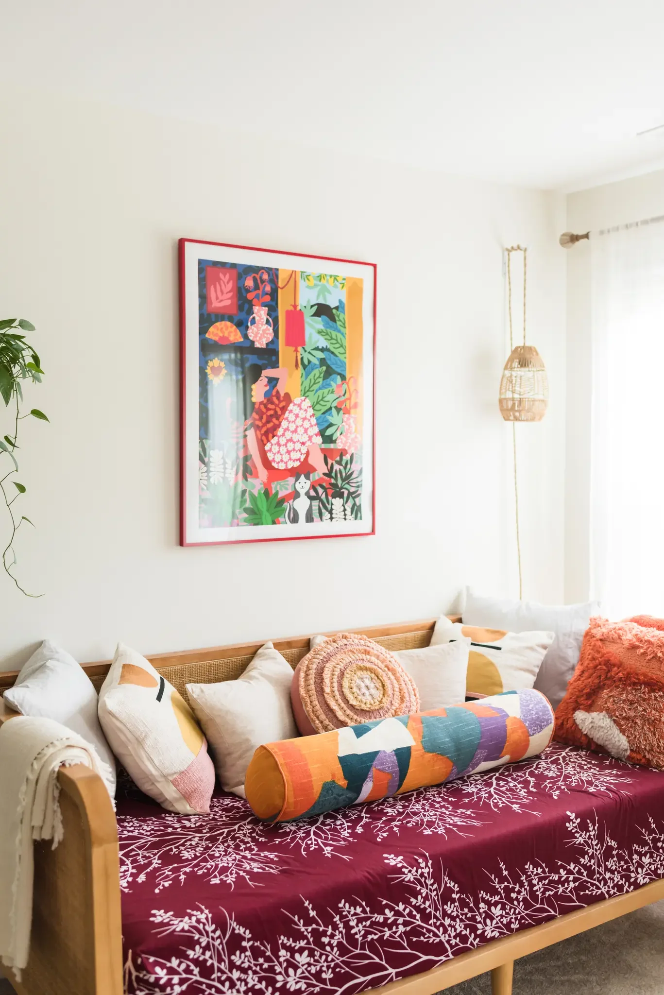 A bedroom with a wooden bed frame, a maroon bedspread with a white leaf and branch pattern, and assorted pillows and throws. Above the bed, colorful artwork in a red frame hangs on a white wall. A woven pendant light fixture hangs from the ceiling to the right. There is a window with sheer curtains and a possible plant on the left.