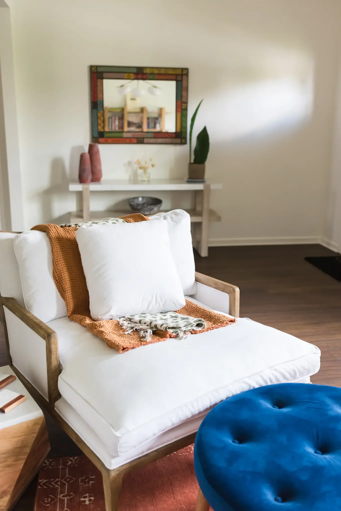 A white bed with pillows and an orange throw blanket, a wooden chair with a blue cushion, a white console table with decorative vases, a mirror on the wall, a tall plant, and bookshelf reflections visible in the mirror, all in a cozy living room.
