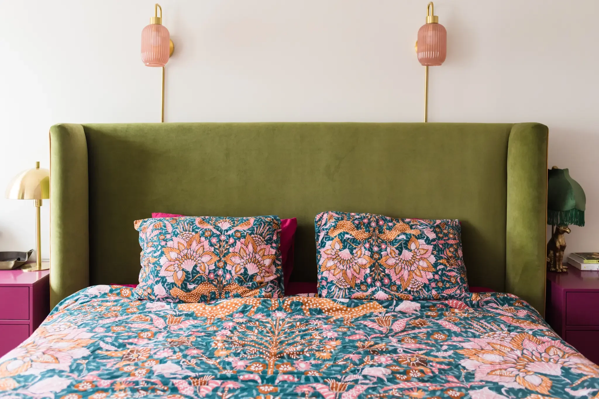 A bedroom with a green velvet headboard, colorful floral bedding, pink bedside tables, and pink wall sconces.