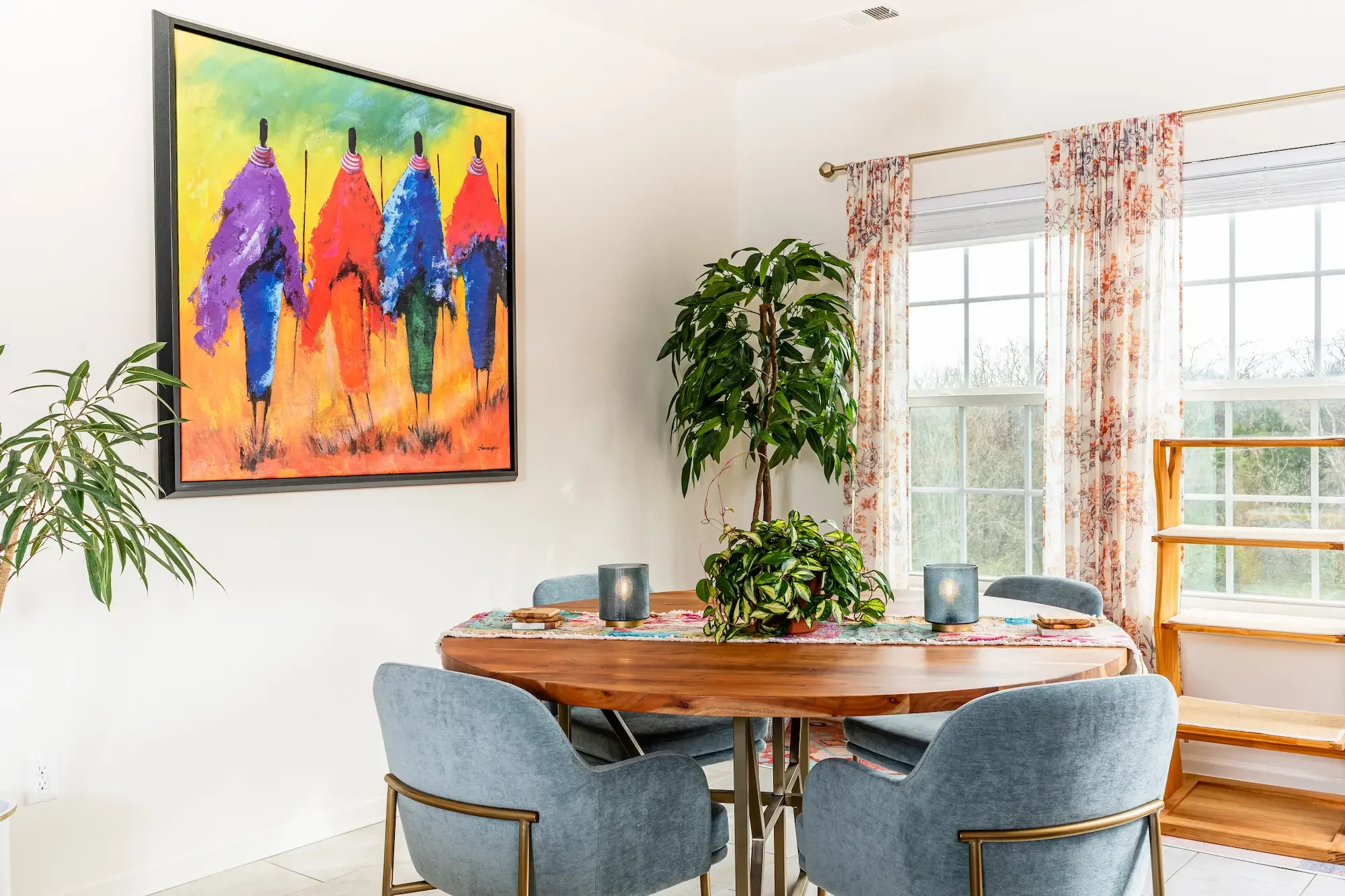 A dining room with a wooden table surrounded by four gray upholstered chairs, a colorful table runner, and two teal candle holders. A large leafy plant is on the table, and several other plants are nearby. The room has white walls, a bright window with floral curtains, and a vibrant colorful painting of abstract figures on the wall.