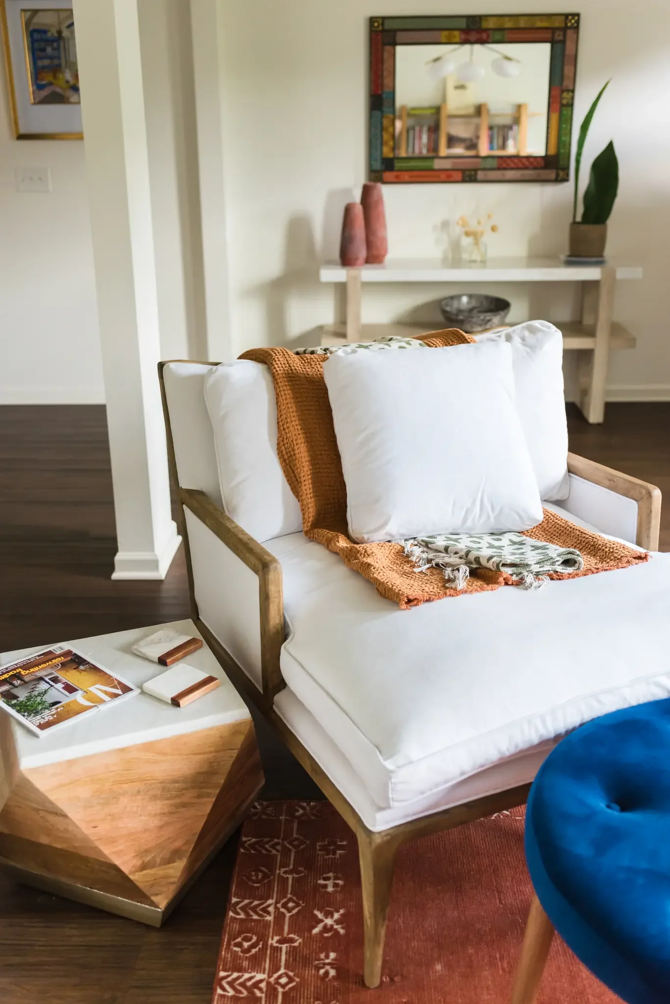 A cozy living room featuring a white cushioned armchair with a wooden frame, draped with an orange knitted blanket. There is a small wooden side table with a magazine, a marble coaster, and some coasters. In the background, a white console table holds two tall reddish-brown vases, a small vase with yellow flowers, and a potted plant. Above, a colorful framed mirror reflects a bookshelf and ceiling light fixture. The room has dark wood flooring and a red patterned area rug.