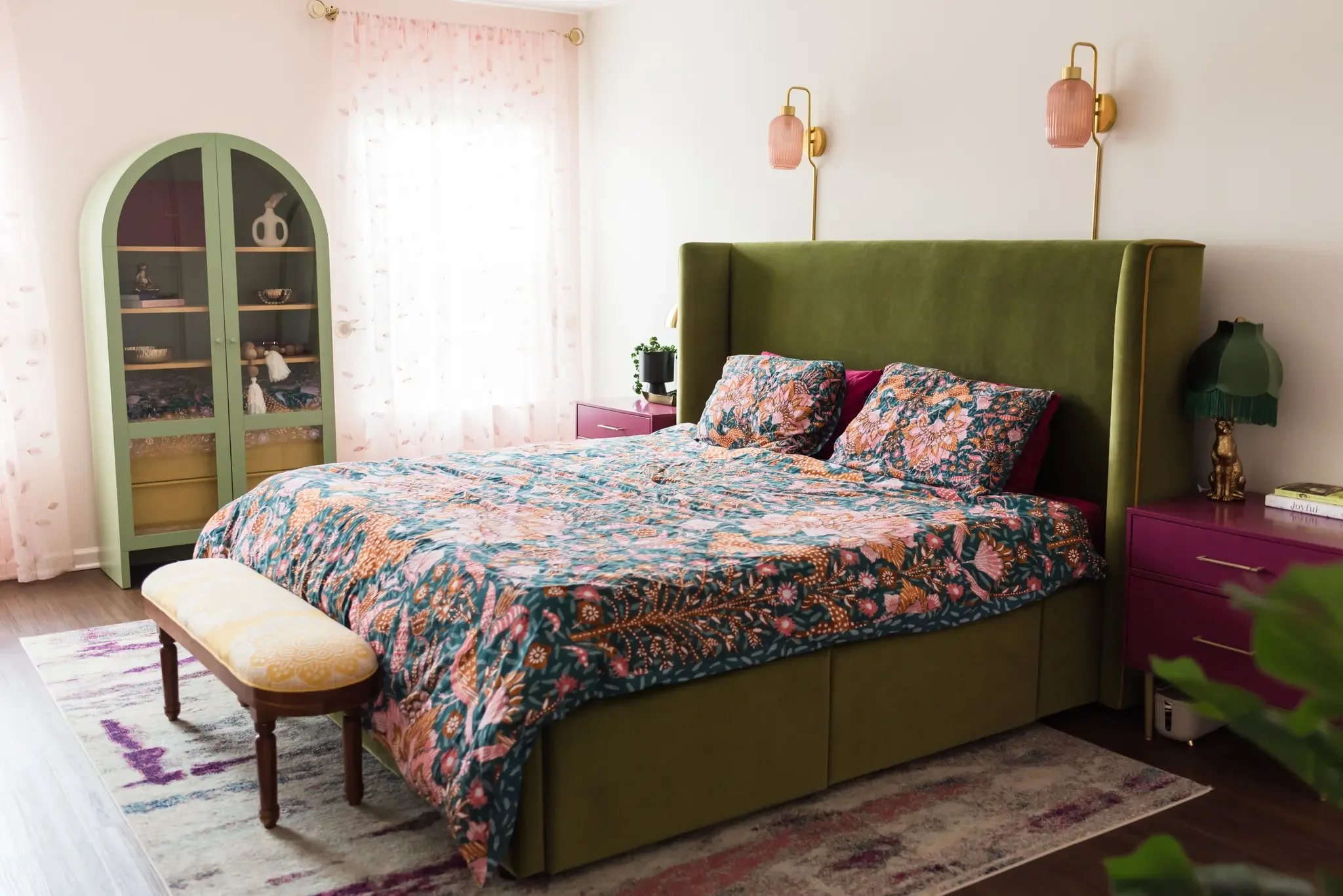 A bedroom with a green upholstered bed, floral bedding, pink nightstand, and matching pink wall sconces. There is a green display cabinet with decorative items and a window with sheer curtains.