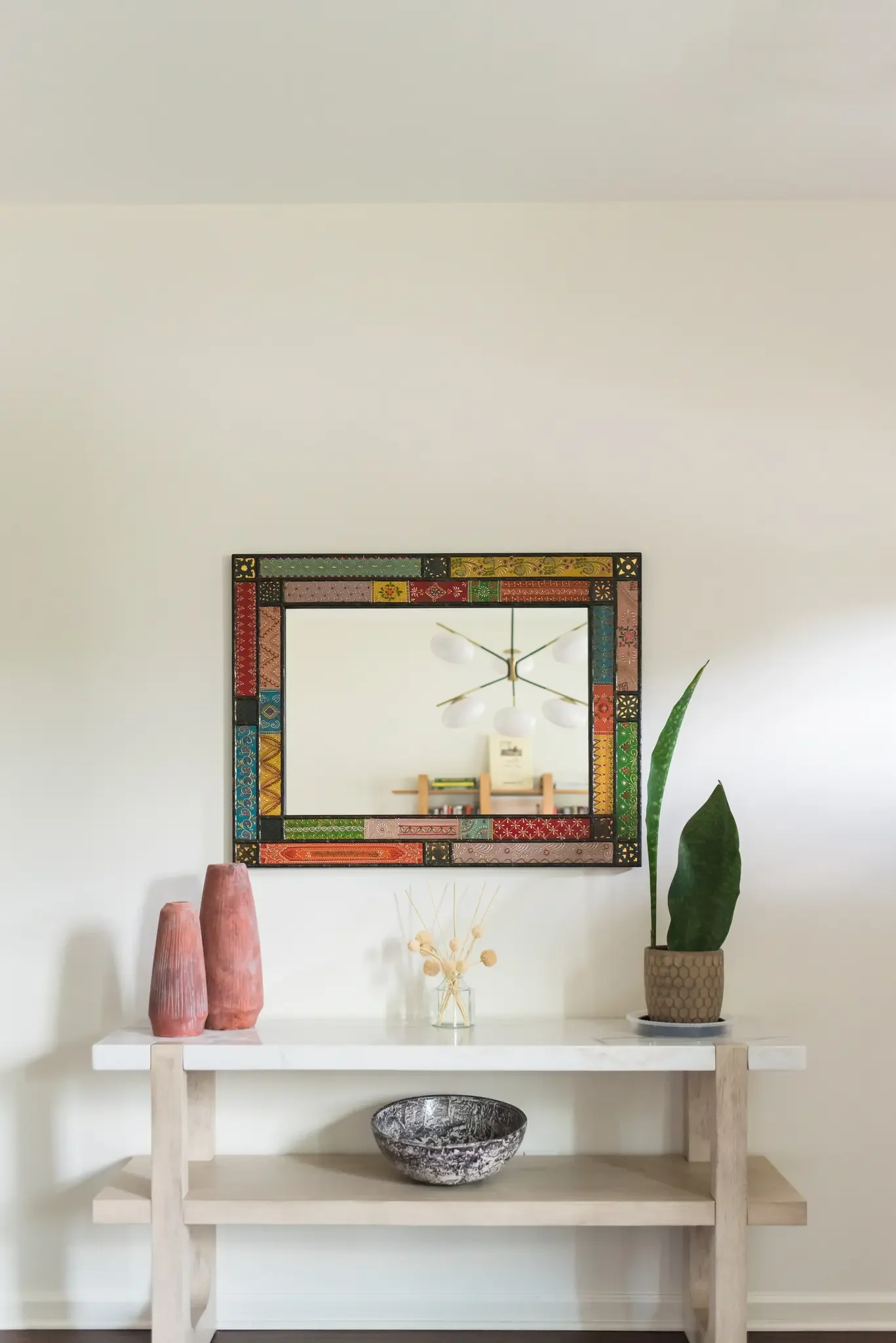 A white console table decorated with two pink vases, a glass jar with dried flowers, and a large bowl. A potted plant is on the right, and a colorful framed mirror hangs above the table. A modern chandelier is reflected in the mirror.