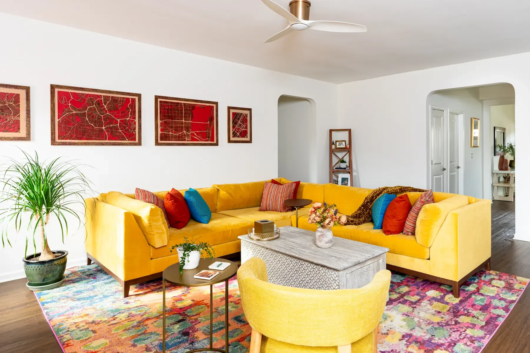 A colorful living room with a bright yellow sectional sofa adorned with red, blue, and striped pillows. A gray coffee table with a floral arrangement and a small round side table with plants and books are in front of the sofa. There is a vibrant multicolored area rug under the furniture. A large potted plant is positioned near the sofa. The walls are decorated with red map artwork, and a corner bookshelf with plants and framed photos is visible. The ceiling has a white fan with brass accents, and the room is well-lit with natural light.