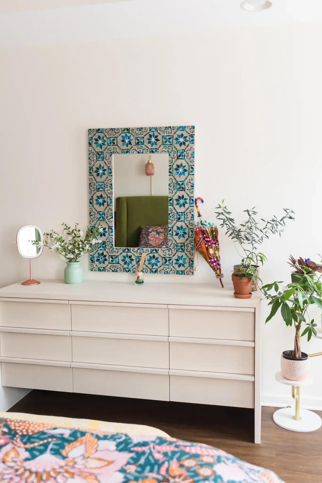 A white dresser with six drawers in a bedroom, decorated with various plants, a mirror, a table lamp, and a colorful umbrella, with a mirror reflecting a green headboard and a wall light.