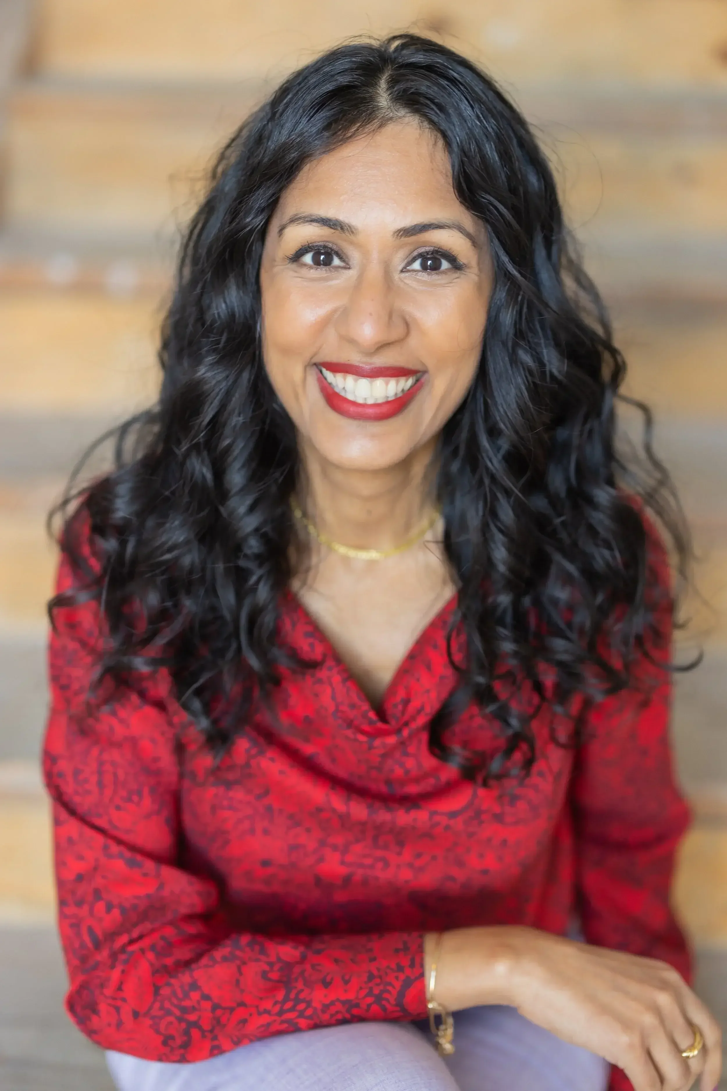 An interior designer with long, curly black hair and brown eyes smiling, wearing a red patterned top, sitting indoors with a wooden wall background.
