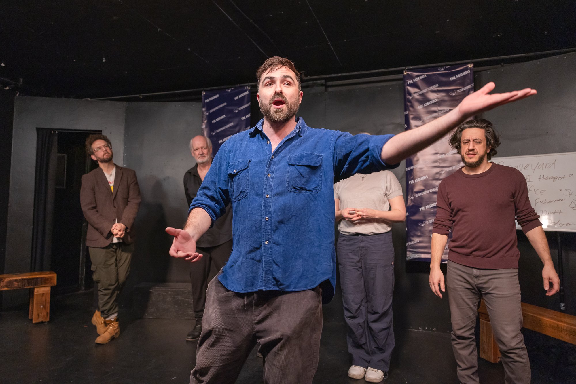 A man with brown hair, a beard, wearing a blue shirt, speaks passionately with arms extended in a black studio space, surrounded by other men listening.