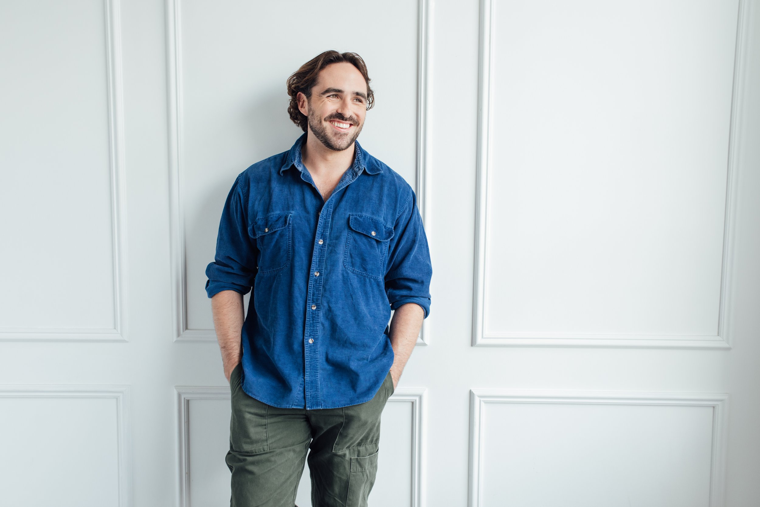 A man with dark hair and a beard smiling, wearing a denim shirt with sleeves rolled up, standing against a white wall with panel molding and gray pants.