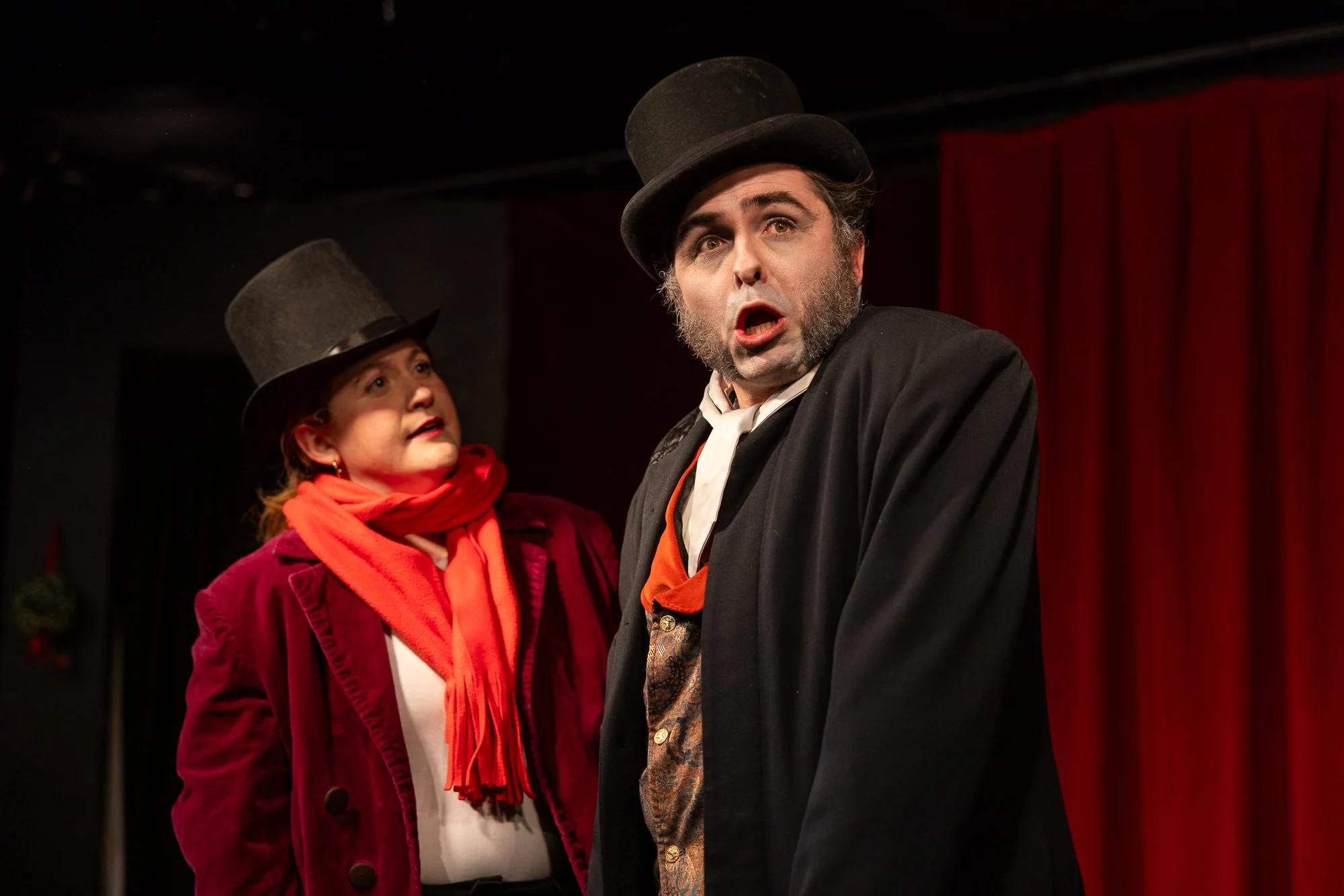 Two actors on stage dressed in vintage clothing, wearing black bowler hats, engaged in a theatrical performance. The man has a surprised expression with his mouth open, and the woman is looking at him attentively. The background features red curtains.