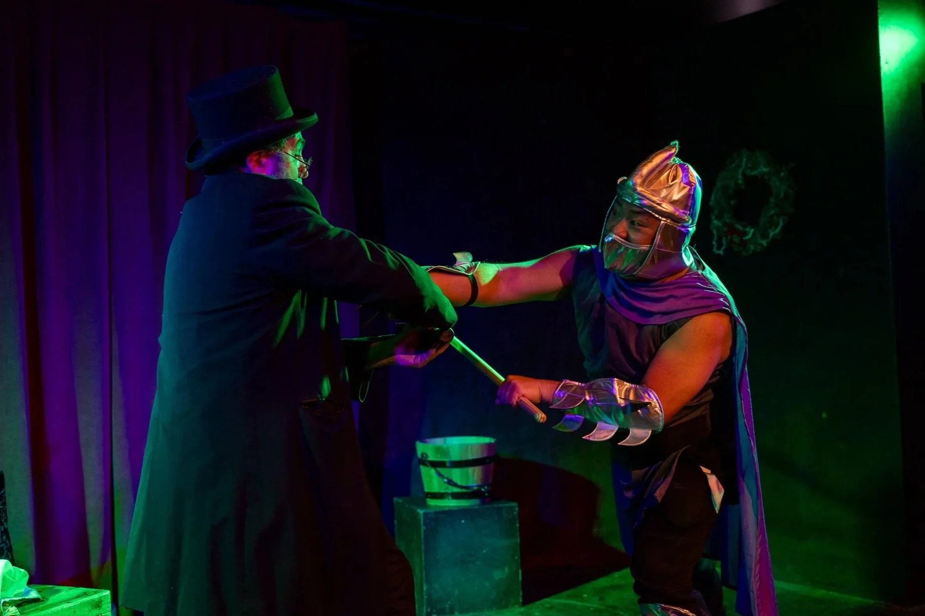 Two performers dressed in costumes engage in a theatrical scene on stage with colorful lighting. One in a top hat and glasses holds a case, while the other, wearing a metallic hat and cape, appears to be acting out a dramatic or fantasy scene.