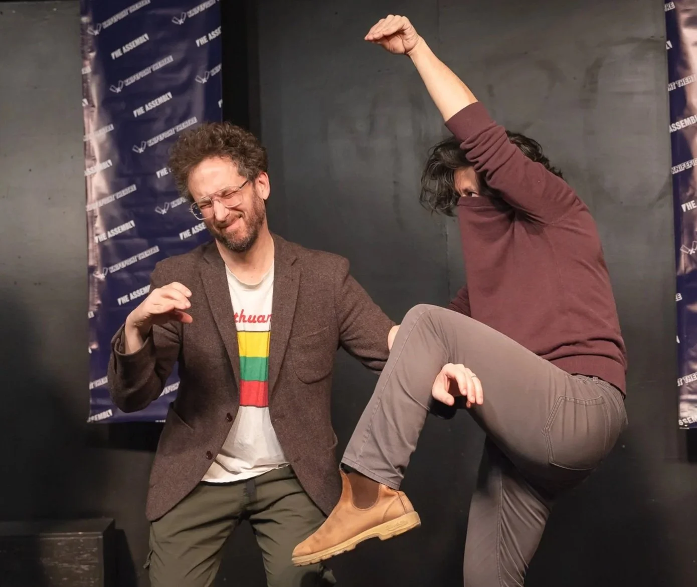 Two men are joking on a stage with a backdrop that says "The Assembly". One man, with glasses and curly hair, is smiling with his eyes closed and has a beard. The other man, with dark curly hair, is raising his arm and has his knee raised, as if in a playful kick, while covering part of his face.