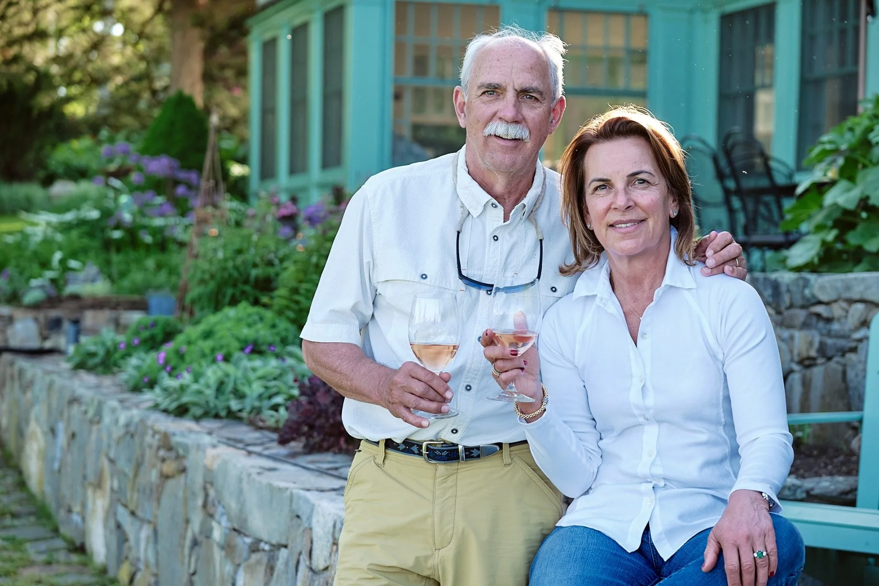 A senior man and woman with wine glasses in a garden, smiling at the camera, with greenery and a glass greenhouse in the background.