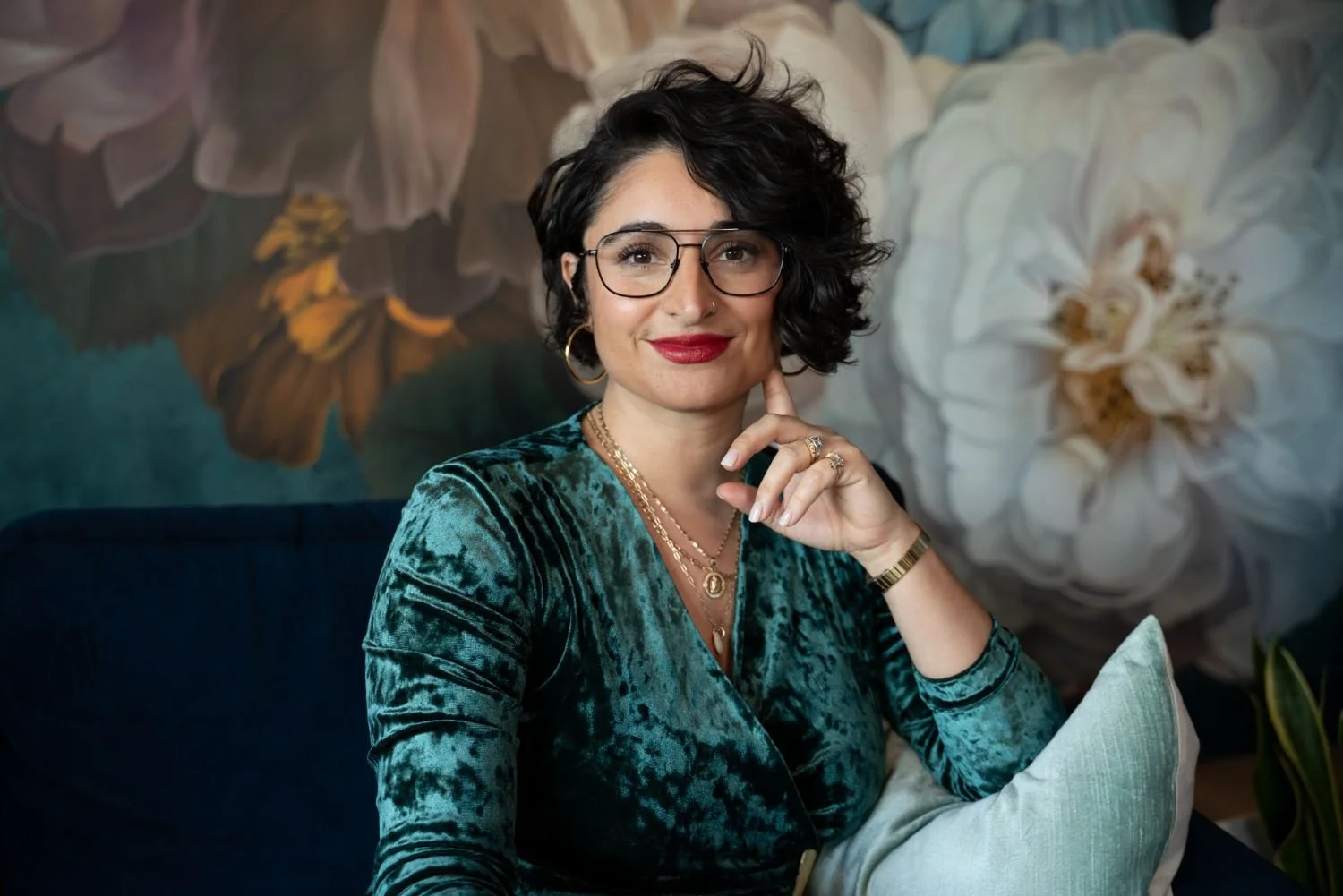 A woman with short, curly black hair, glasses, and red lipstick, sitting on a dark blue sofa with cream cushions, in front of a floral wall mural, wearing a teal velvet top and gold jewelry.
