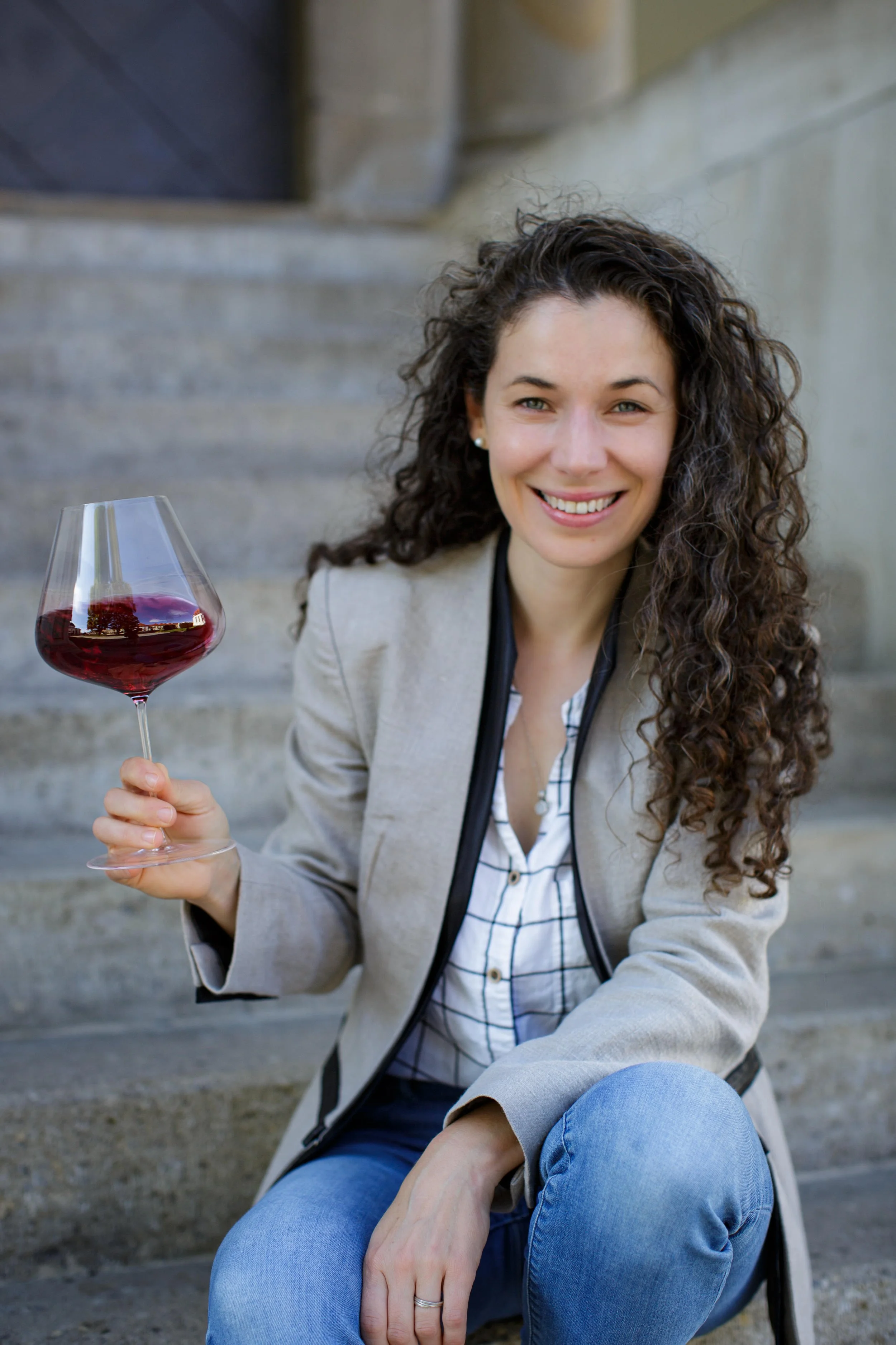 A smiling woman with curly brown hair seated on outdoor stairs, holding a glass of red wine.