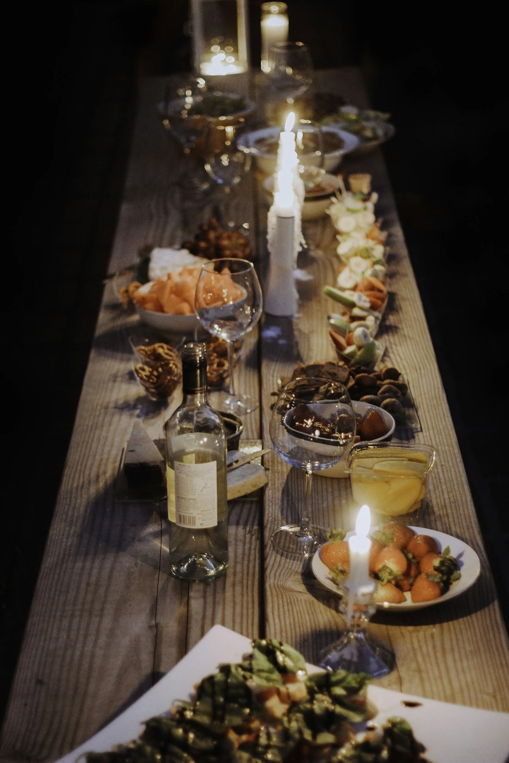 A dinner table with various dishes, bowls of food, wine glasses, and lit candles in a dimly lit setting.