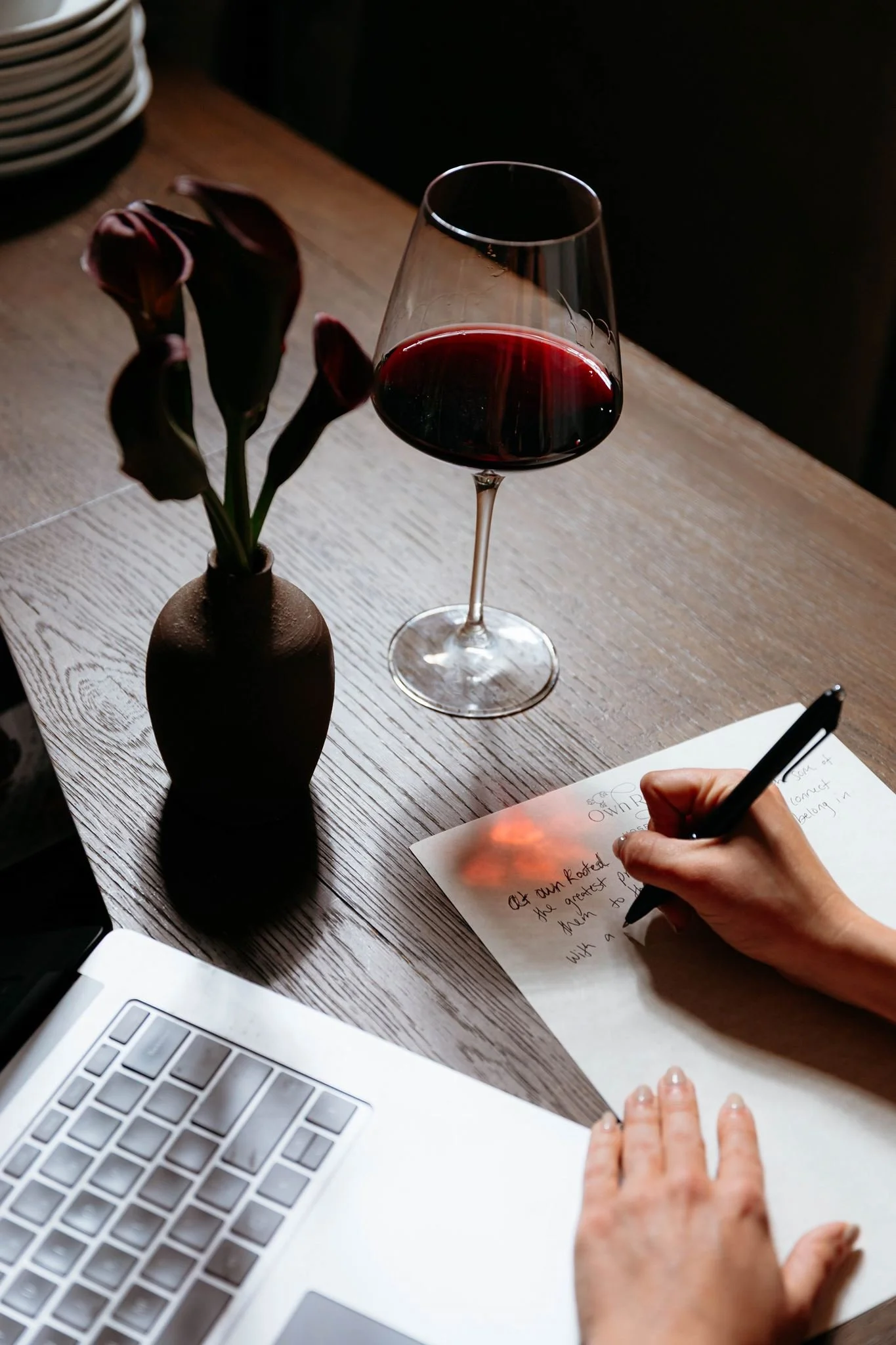 A person is writing notes on a piece of paper on a wooden table. Next to the paper, there's a laptop with a silver keyboard, and a glass of red wine. In the background, there is a vase with dark red tulips and a stack of white plates. The scene is lit with natural light.