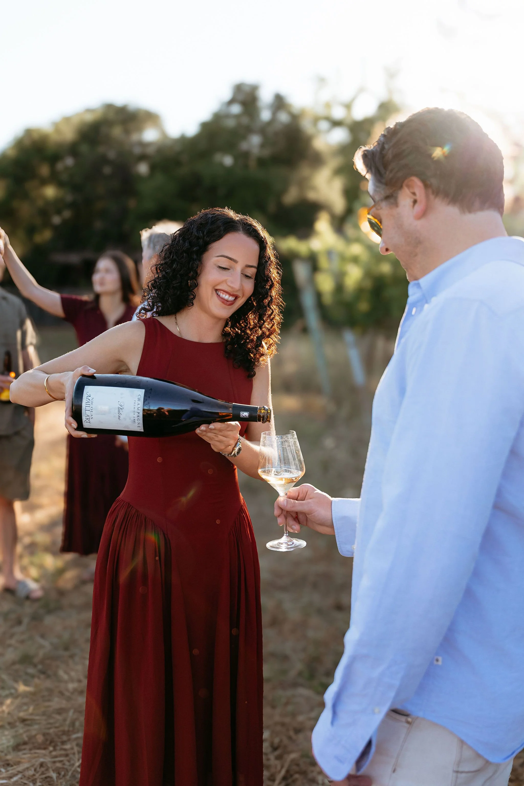 A woman in a red dress pouring champagne into a glass held by a man in a pastel blue shirt during an outdoor gathering with several people and trees in the background.