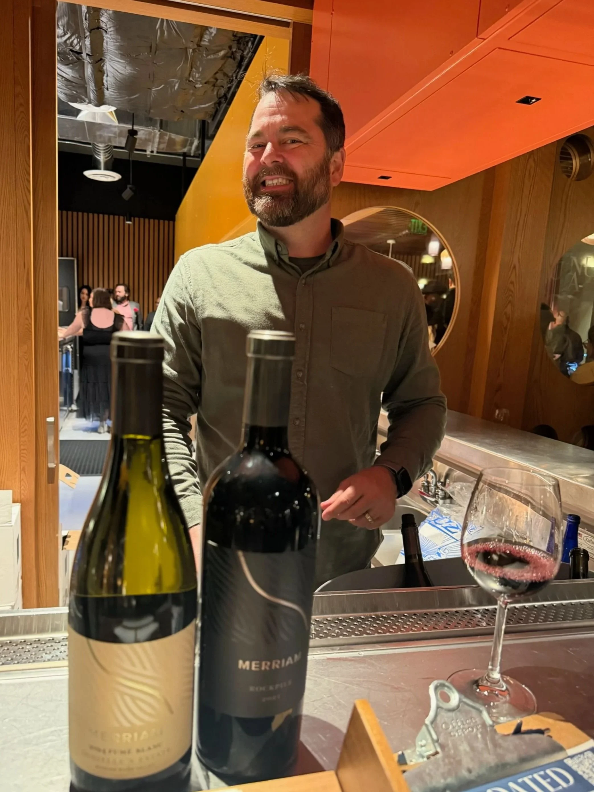 A man with a beard smiling at a wine tasting event with bottles of wine, glasses, and wine openers on the counter, and a warm, modern interior with wooden accents and other guests in the background.