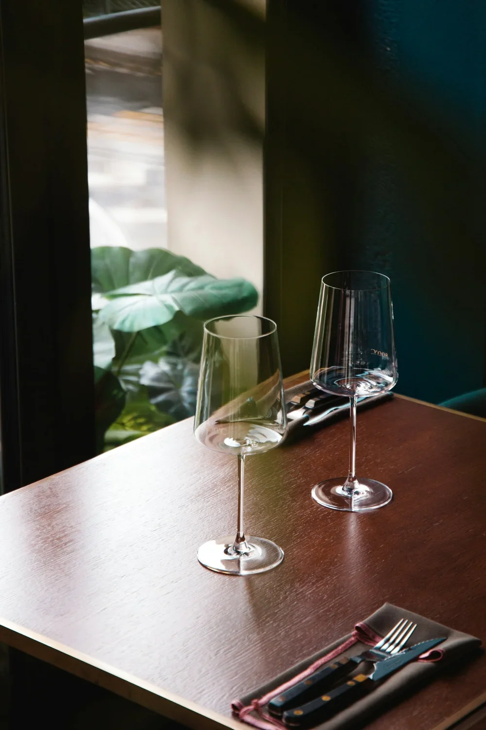 Two empty wine glasses on a wooden table set for dining, with a cloth napkin and cutlery on the corner, near a window showing some foliage outside.