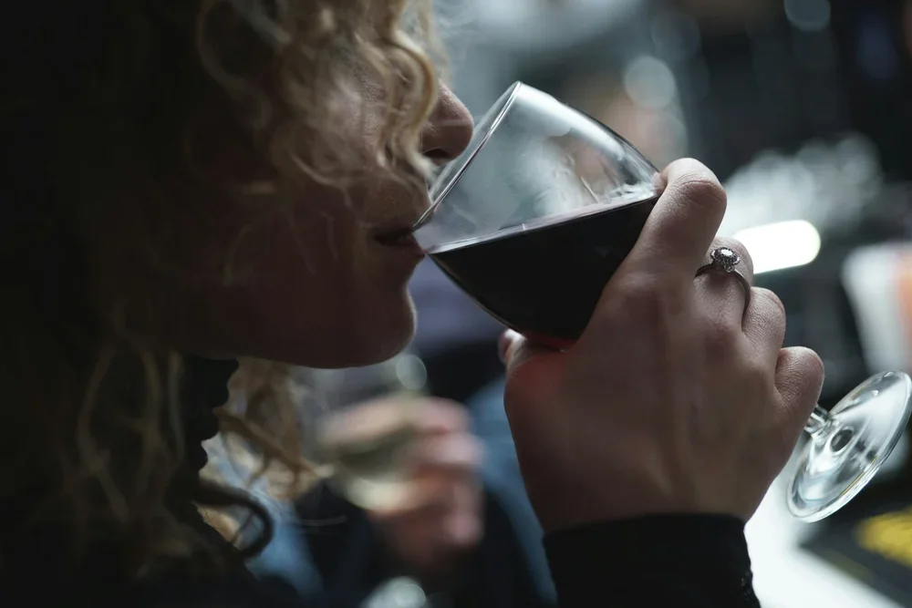 A woman with curly hair drinking red wine from a glass in a dimly lit setting.